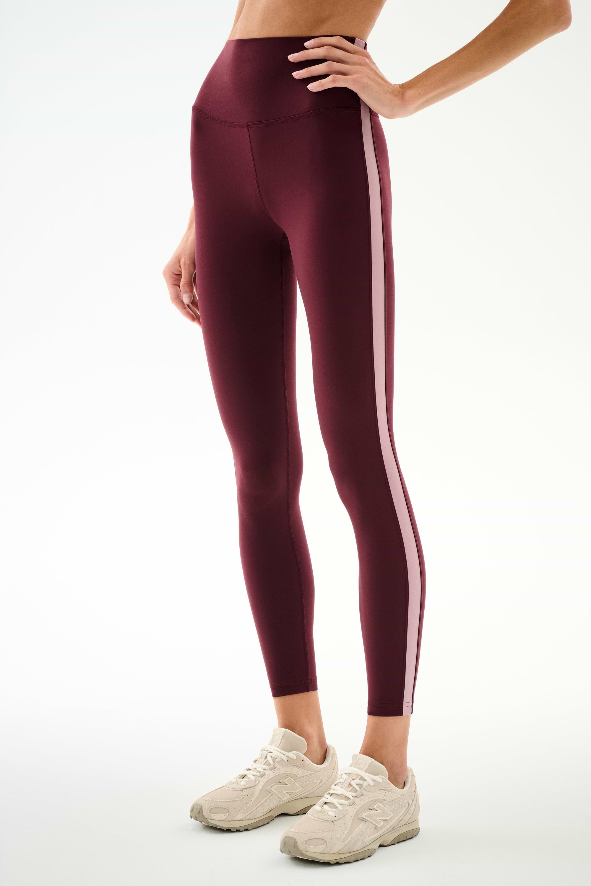 A person is wearing the SPLITS59 Clare High Waist Rigor 7/8 leggings in Dark Cherry/Blush, featuring a high-waisted sculpting fit with light pink side stripes and paired with beige sneakers, standing against a plain white background. These workout leggings combine comfort and style.