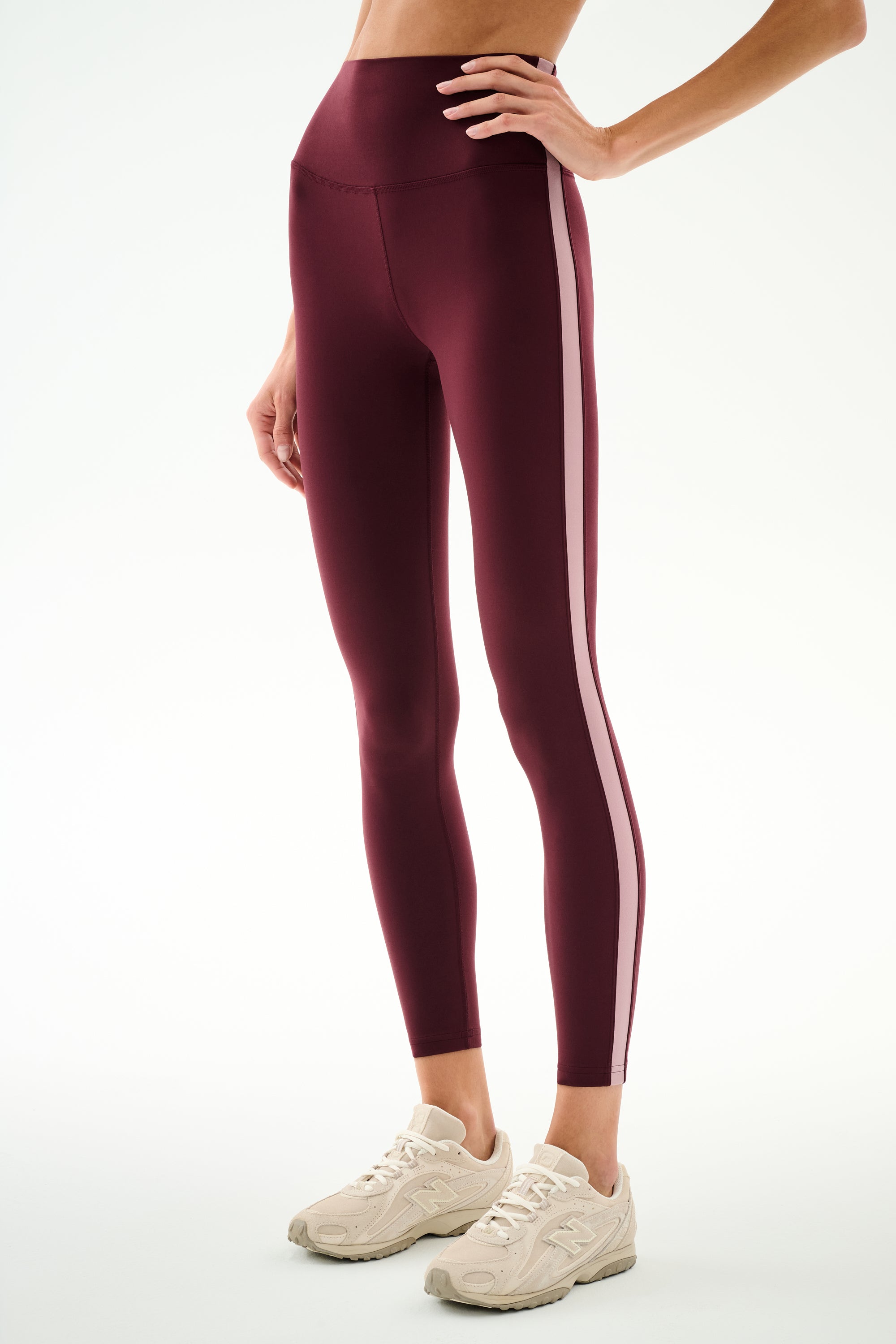 A person is wearing the SPLITS59 Clare High Waist Rigor 7/8 leggings in Dark Cherry/Blush, featuring a high-waisted sculpting fit with light pink side stripes and paired with beige sneakers, standing against a plain white background. These workout leggings combine comfort and style.