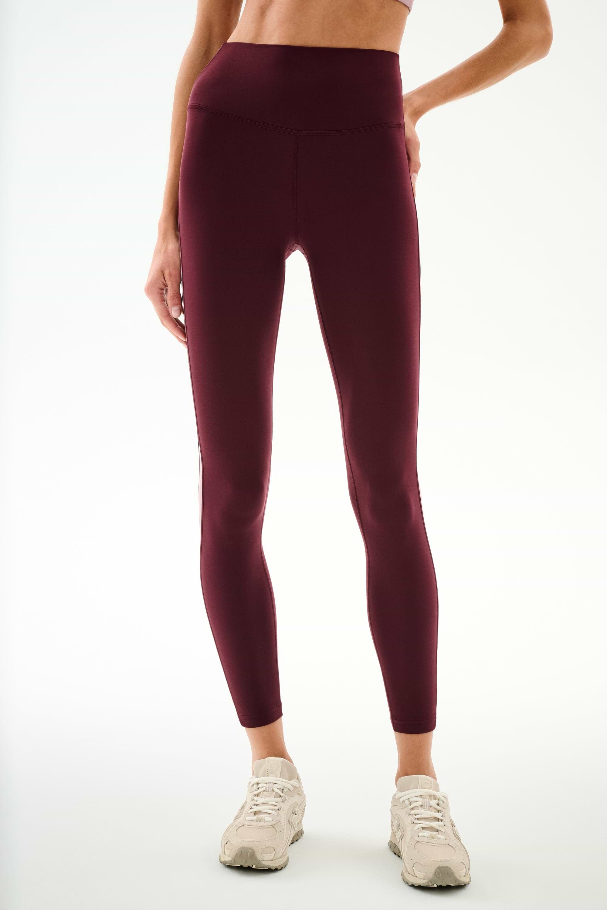 A person is wearing SPLITS59 Clare High Waist Rigor 7/8 leggings in Dark Cherry/Blush, featuring a sculpting high-waisted fit, paired with white sneakers and standing against a plain light background.