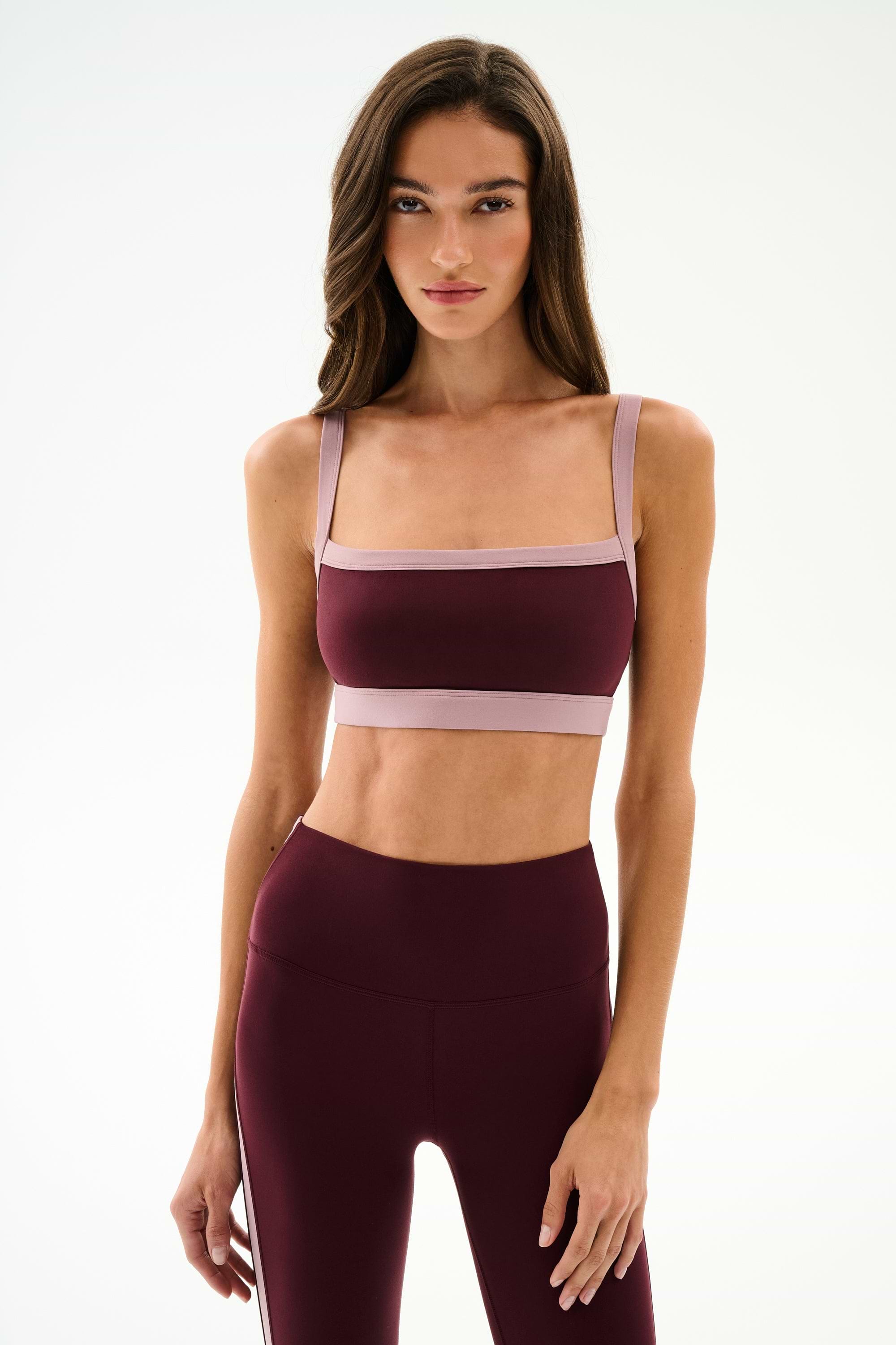 A woman wearing the SPLITS59 Monah Rigor Bra in Dark Cherry/Blush with coordinating high-waisted leggings, standing against a plain white background.