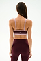 A woman stands with her back to the camera, wearing the SPLITS59 Monah Rigor Bra in Dark Cherry/Blush with wide straps, paired with burgundy high-waisted leggings, against a plain light background.