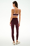 A woman stands with her back to the camera, wearing the SPLITS59 Monah Rigor Bra in Dark Cherry/Blush, paired with maroon leggings and white sneakers against a plain, light background.