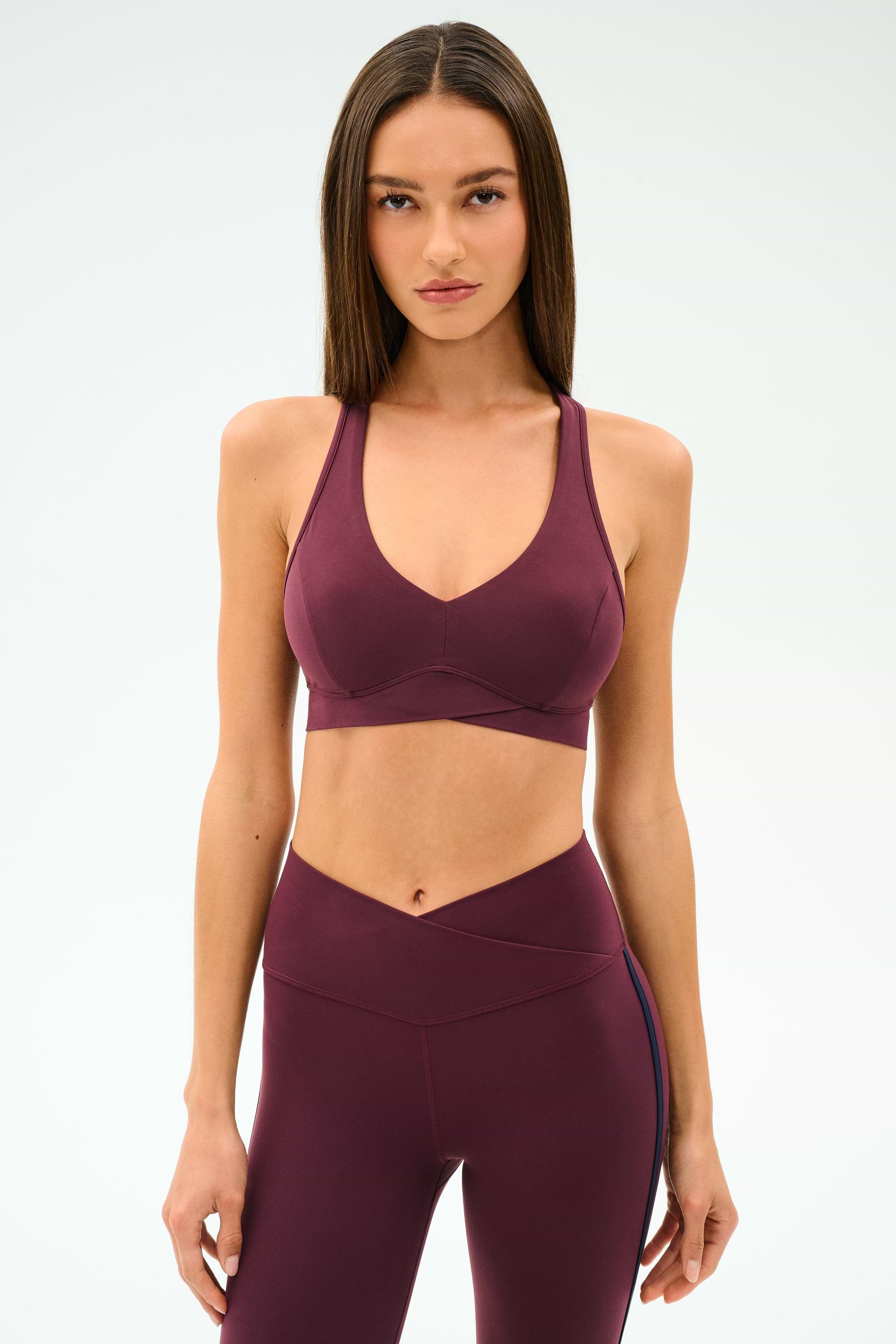 Woman stands against a plain background, wearing the SPLITS59 Mia Rigor Bra in Dark Cherry paired with matching high-waisted leggings.