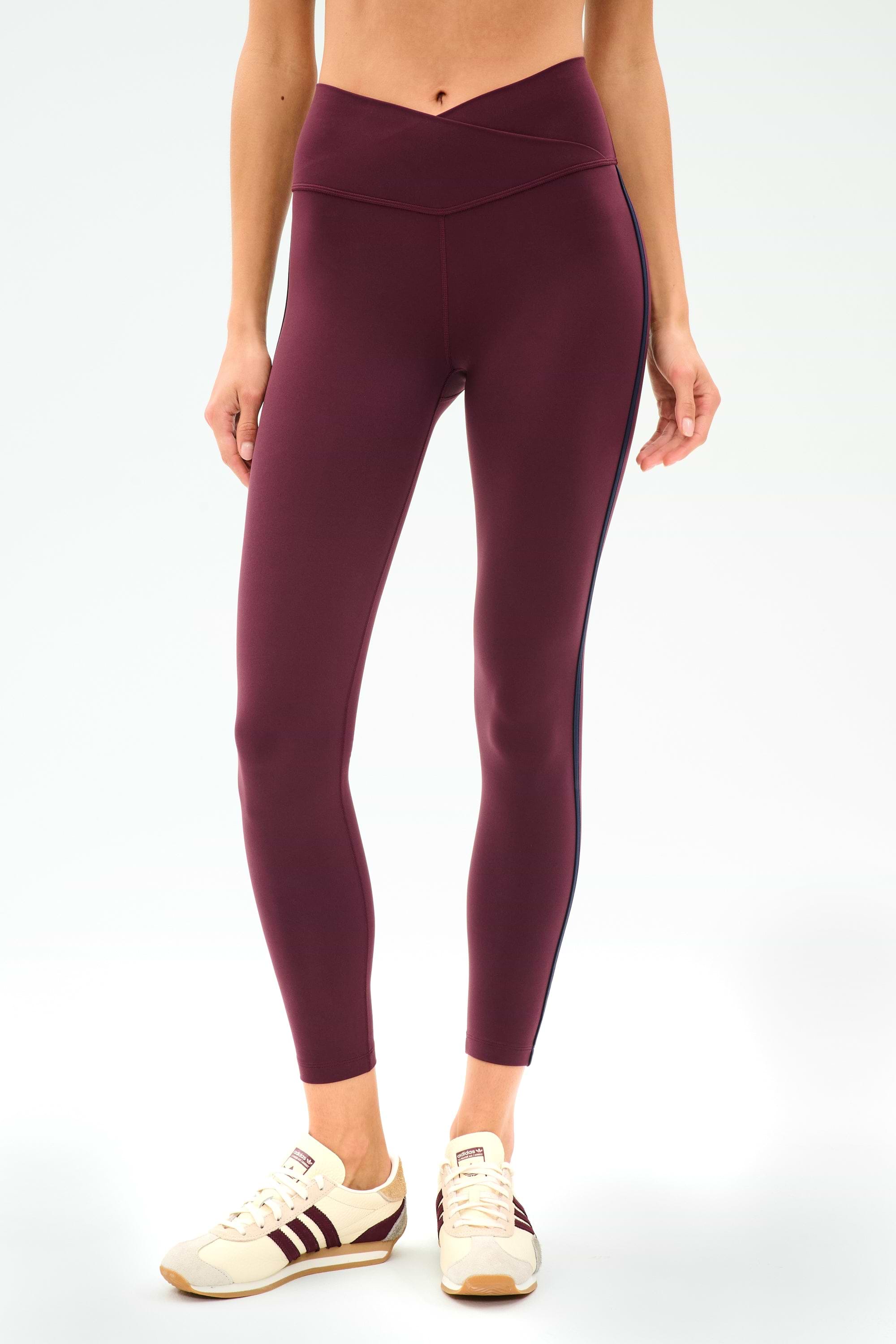 A person wearing SPLITS59 Mia Rigor Legging w/ Stripe in Dark Cherry/Indigo, featuring a V-shaped waistband, paired with beige sneakers with maroon stripes, stands against a plain white background.