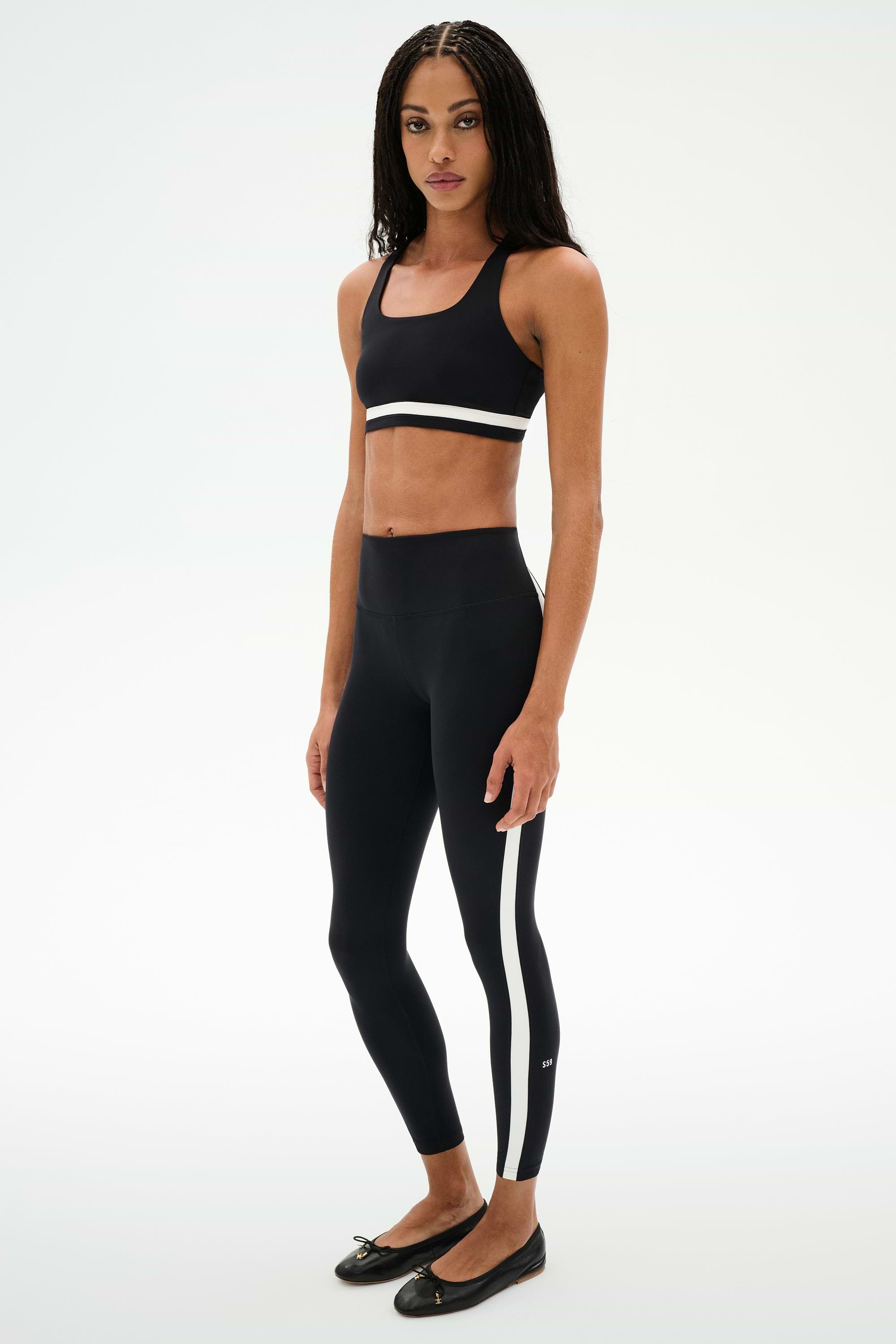 A woman stands in a studio wearing a black sports bra and SPLITS59 Miles High Waist Rigor 7/8 leggings in Black/White, paired with black ballet flats.