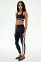 A woman stands in a studio wearing a black sports bra and SPLITS59 Miles High Waist Rigor 7/8 leggings in Black/White, paired with black ballet flats.
