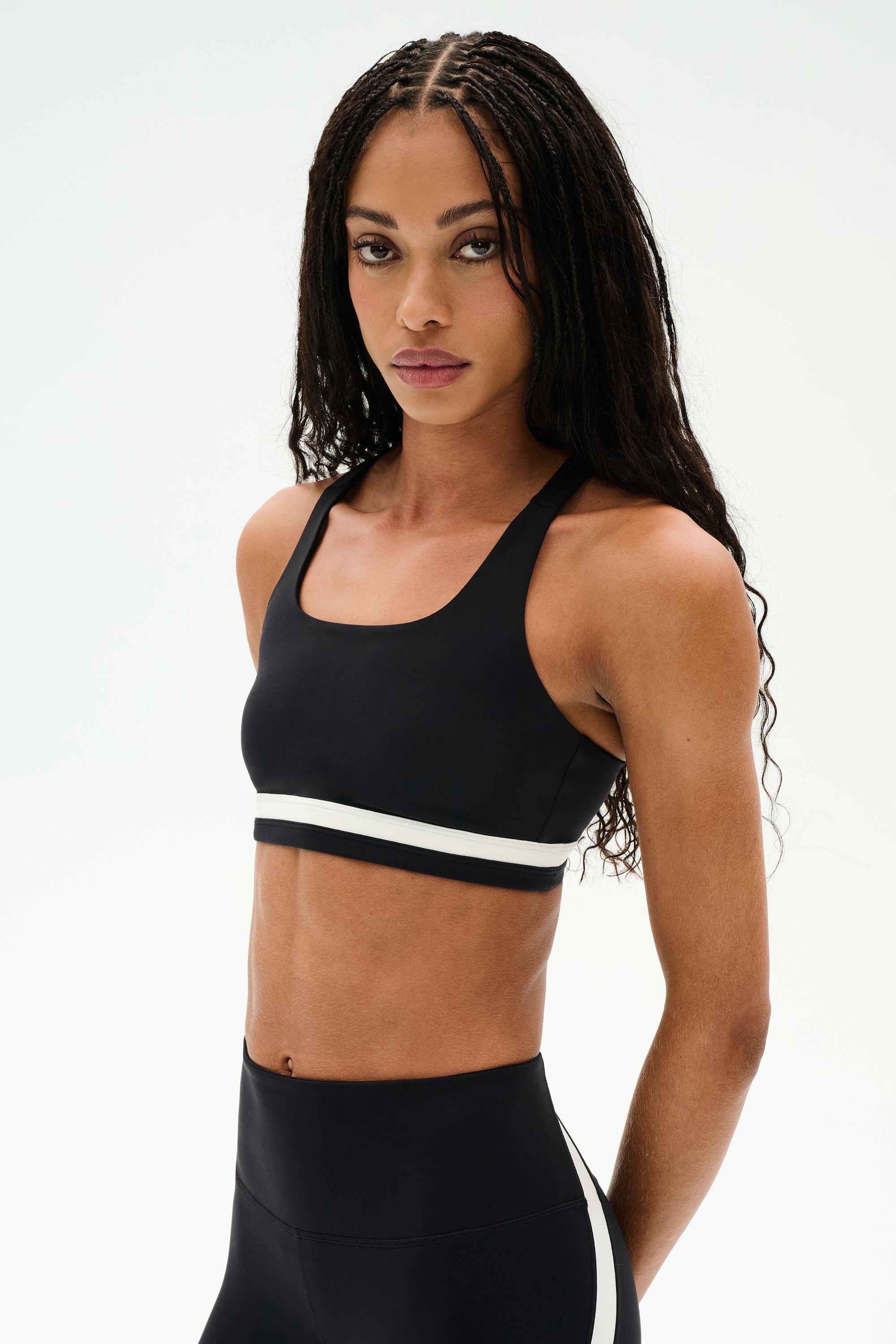 A woman with long braided hair models the SPLITS59 Miles Rigor Bra - Black/White and matching high-waisted leggings against a plain white background, highlighting stylish workout support for women.