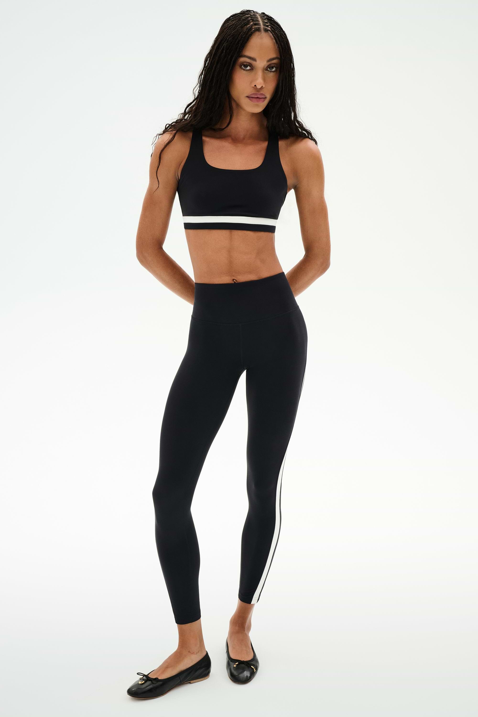 A woman faces forward, wearing SPLITS59's Miles High Waist Rigor 7/8 leggings in Black/White and a black sports bra. She completes the look with black ballet flats.