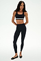 A woman faces forward, wearing SPLITS59's Miles High Waist Rigor 7/8 leggings in Black/White and a black sports bra. She completes the look with black ballet flats.