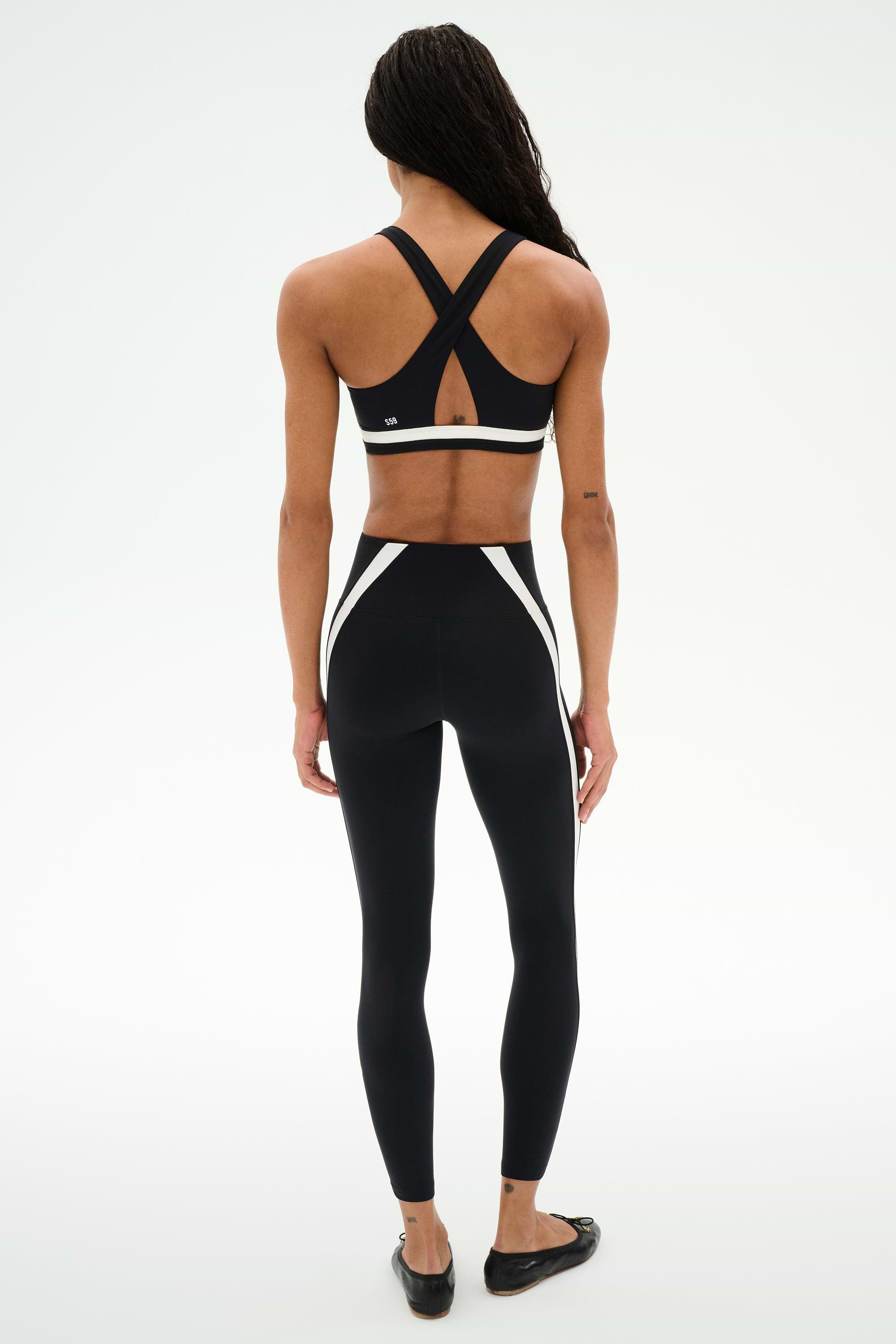 A person faces away, wearing SPLITS59’s Miles High Waist Rigor 7/8 leggings in Black/White with a black crisscross sports bra, set against a plain white background.