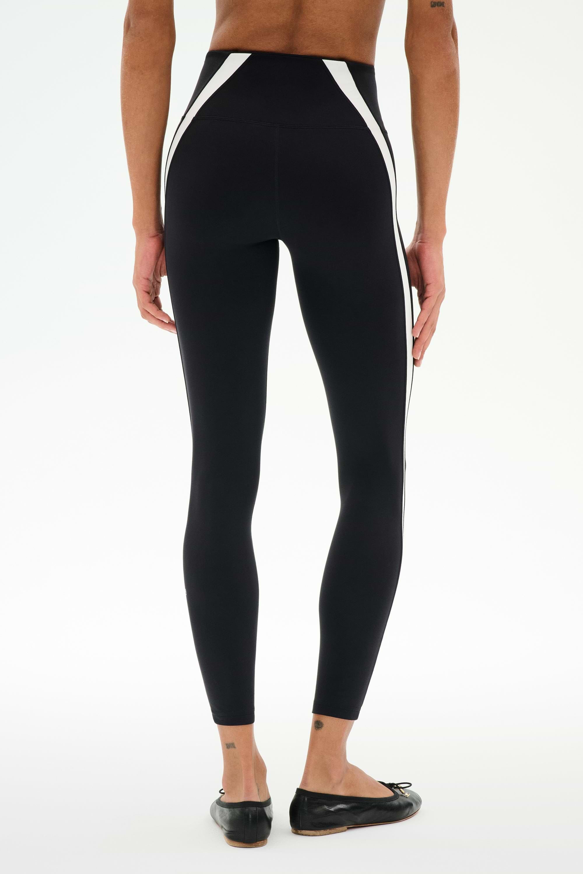 Model wears SPLITS59 Miles High Waist Rigor 7/8 leggings in Black/White, featuring white side panels and black flat shoes, standing with back to camera against a plain white background.