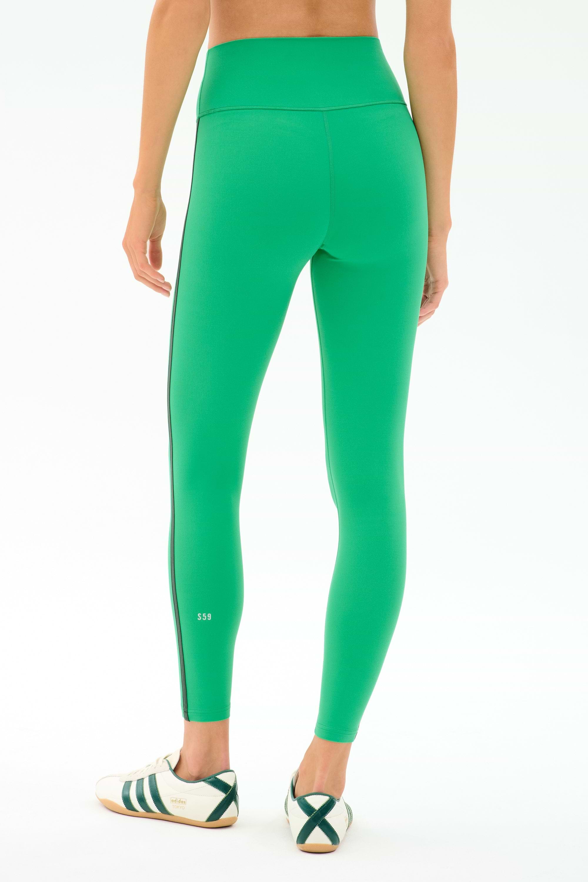 A person wearing SPLITS59 Amber Airweight High Waist 7/8 leggings in Spearmint/Military, paired with white sneakers featuring green stripes, stands on a plain white background, shown from the back.