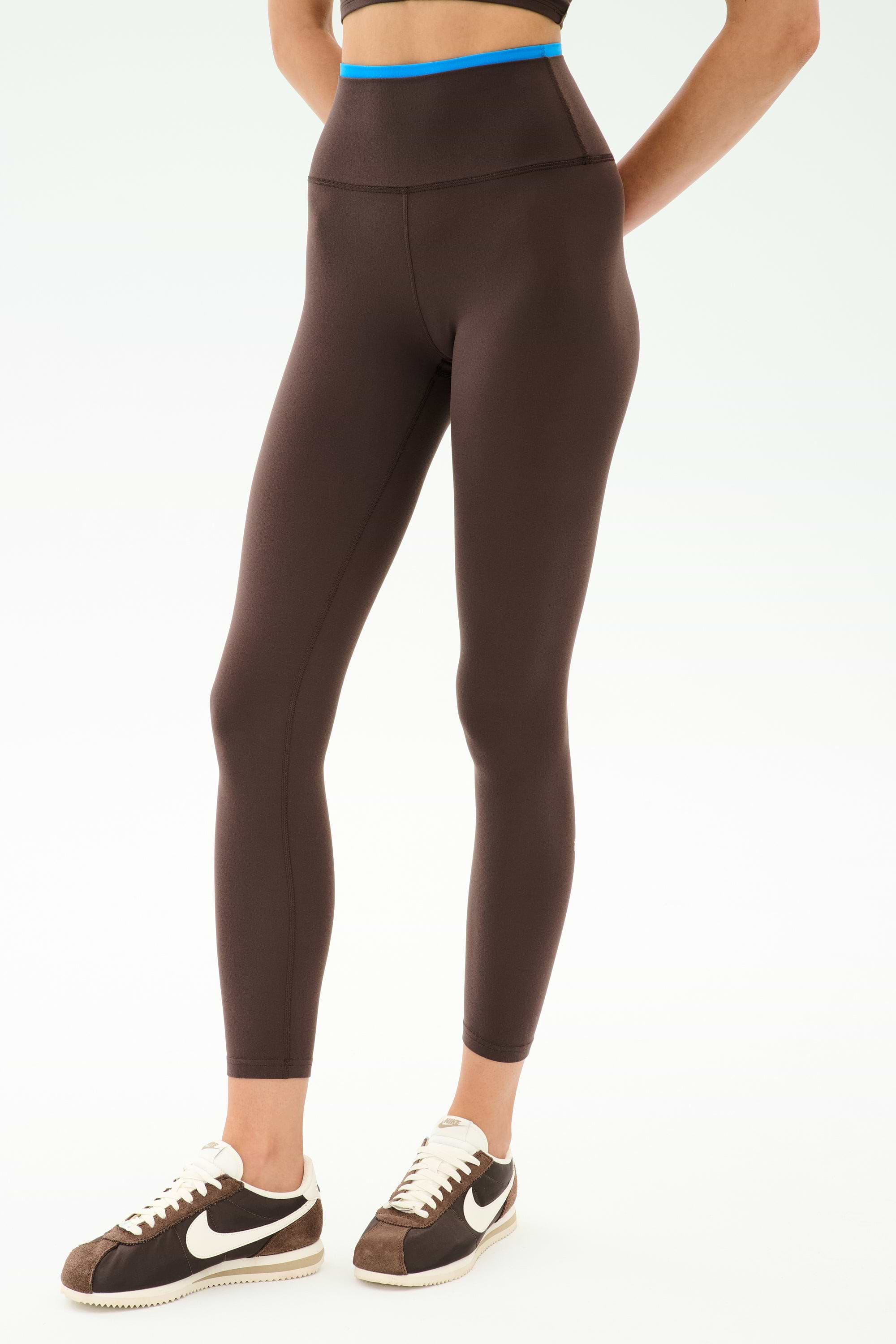 A person wears SPLITS59 Dual High Waist Airweight 7/8 leggings in Dark Chocolate/Classic Blue, featuring a blue waistband, paired with brown Nike sneakers, standing against a plain white background.