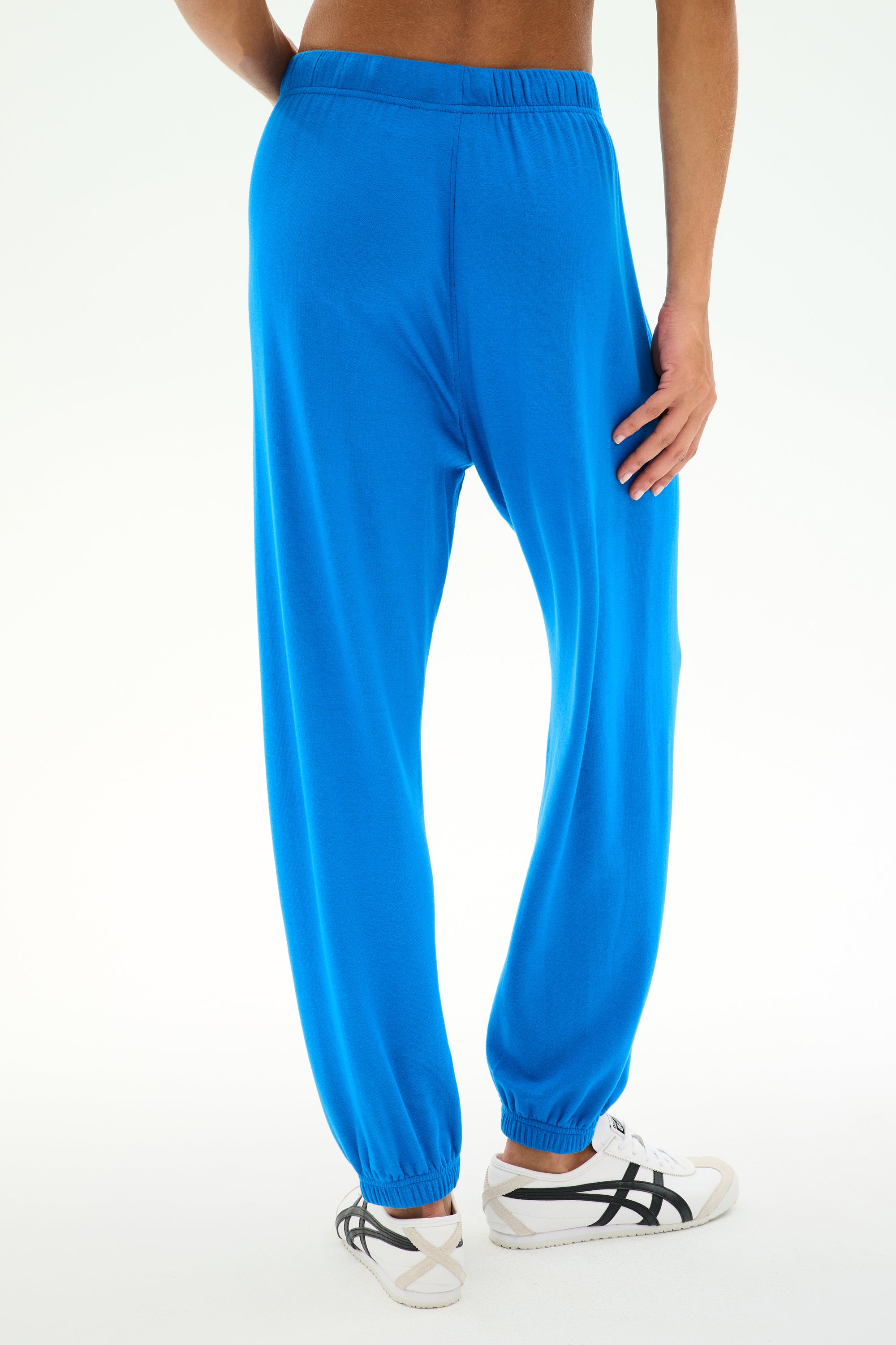 A person wearing the SPLITS59 Andie Oversized Fleece Sweatpant in Classic Blue and white sandals stands with one hand on their hip, shown from the back against a plain background. Made in Los Angeles, these baggy fleece sweatpants combine comfort and style—perfect for cool weather workouts.