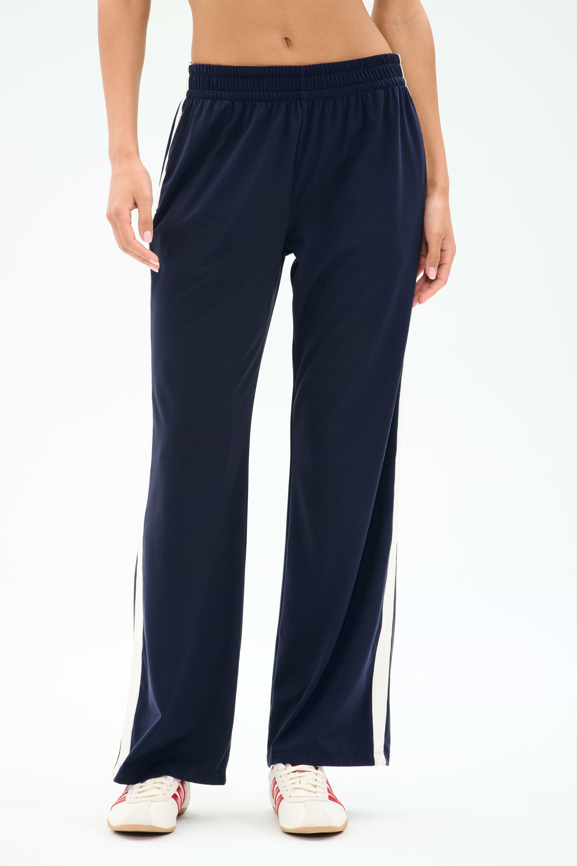 Model wears SPLITS59 Maxie Rigor Track Pant in Indigo/White—navy rigor fabric with white side stripes—paired with white sneakers, shown waist to feet against a plain background.