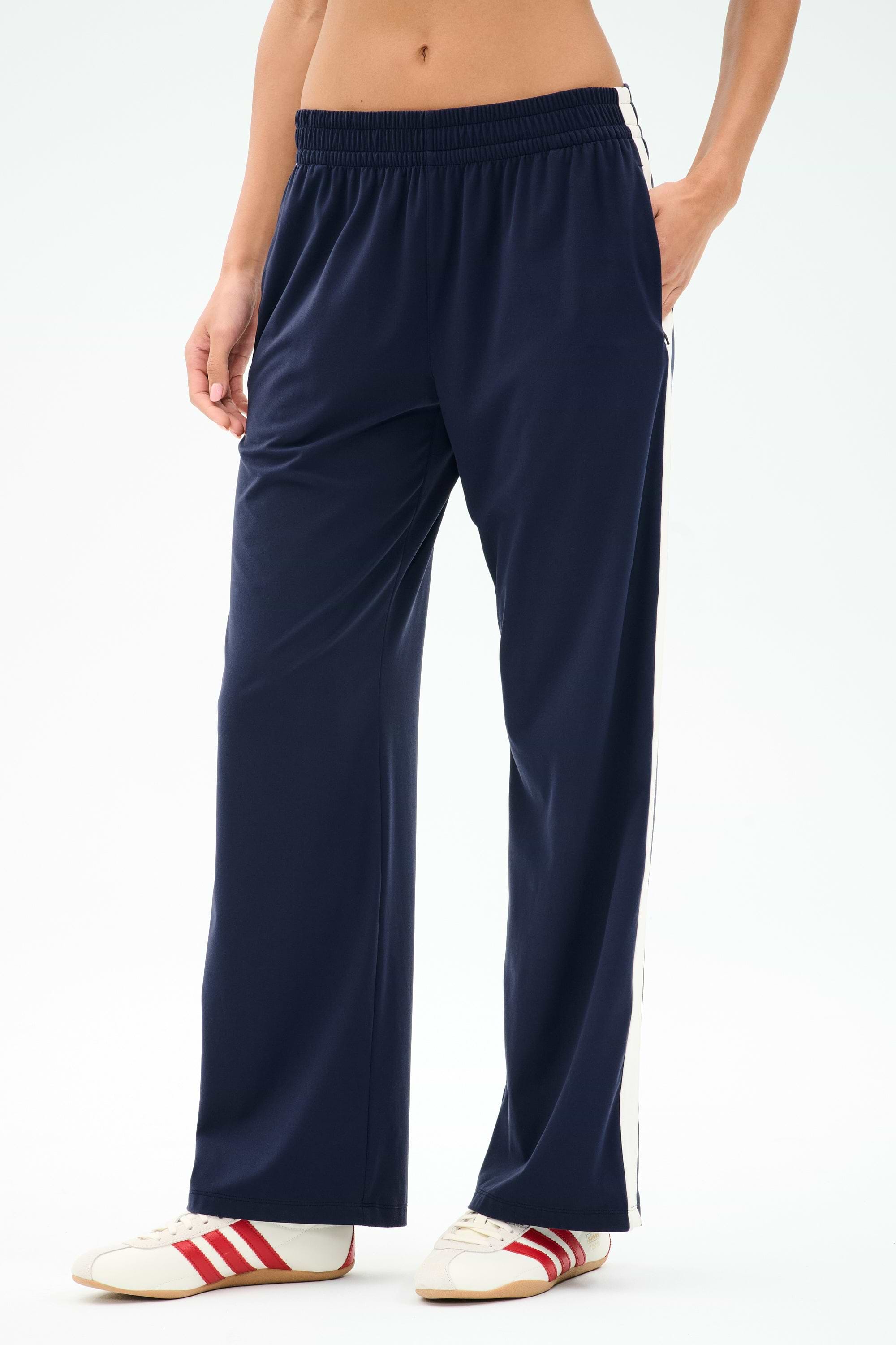 A person wearing SPLITS59 Maxie Rigor Track Pant in Indigo/White with white sneakers featuring red stripes stands against a plain background.