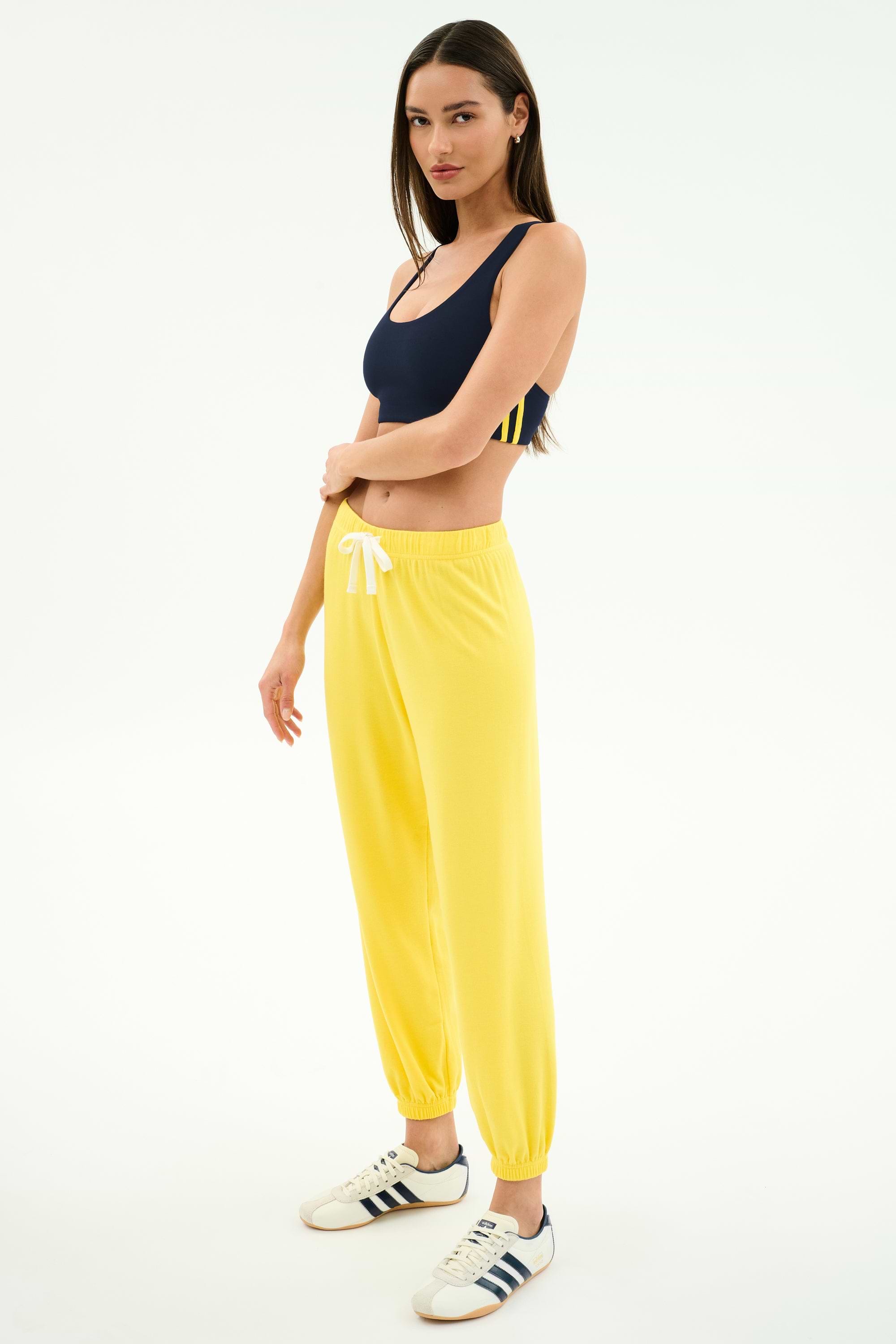 A woman stands in a studio wearing a navy spandex blend sports bra, SPLITS59 Andie Oversized Fleece Sweatpant in bright yellow with a white drawstring, and white Adidas sneakers.