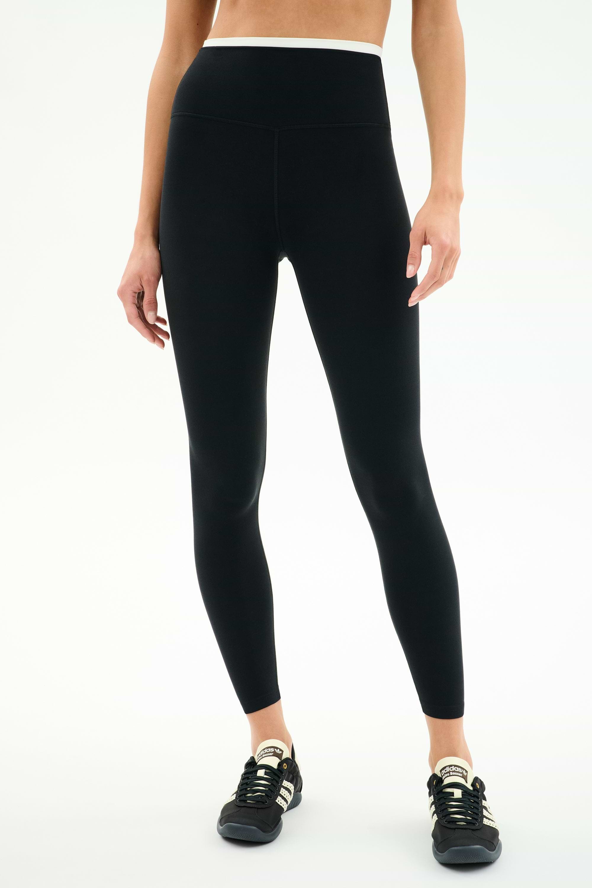 A person wears SPLITS59 Dual High Waist Airweight 7/8 leggings in Black/White, paired with black athletic shoes, standing against a plain white background.