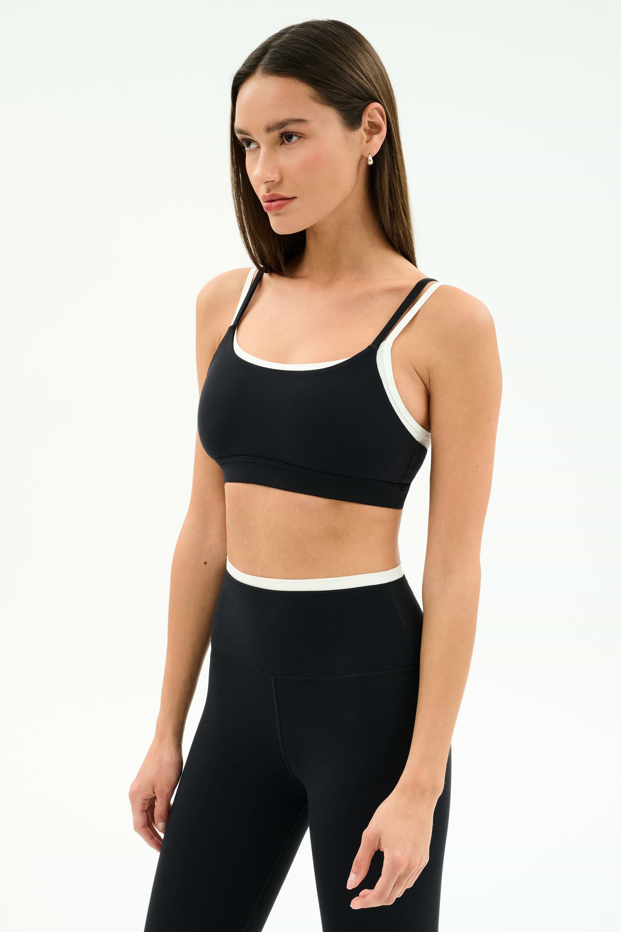 A woman wears the SPLITS59 Elliah Airweight Double Layer Bra in Black/White and high-waisted leggings, posing against a plain white background.