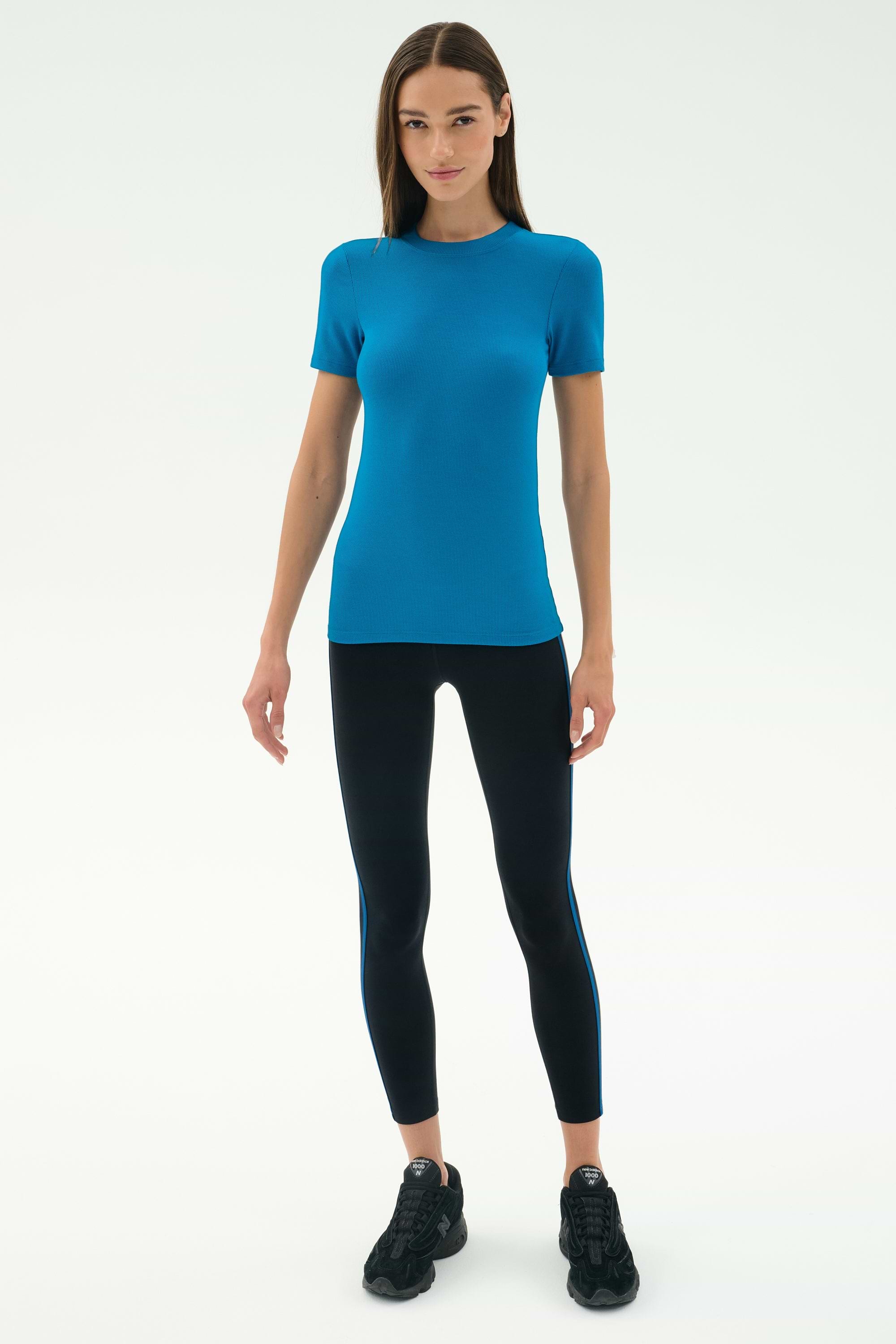 A woman stands facing forward wearing the SPLITS59 Louise Rib Short Sleeve in Stone Blue, black leggings with blue side stripes, and black athletic shoes—perfect for yoga or gym workouts against a white background.