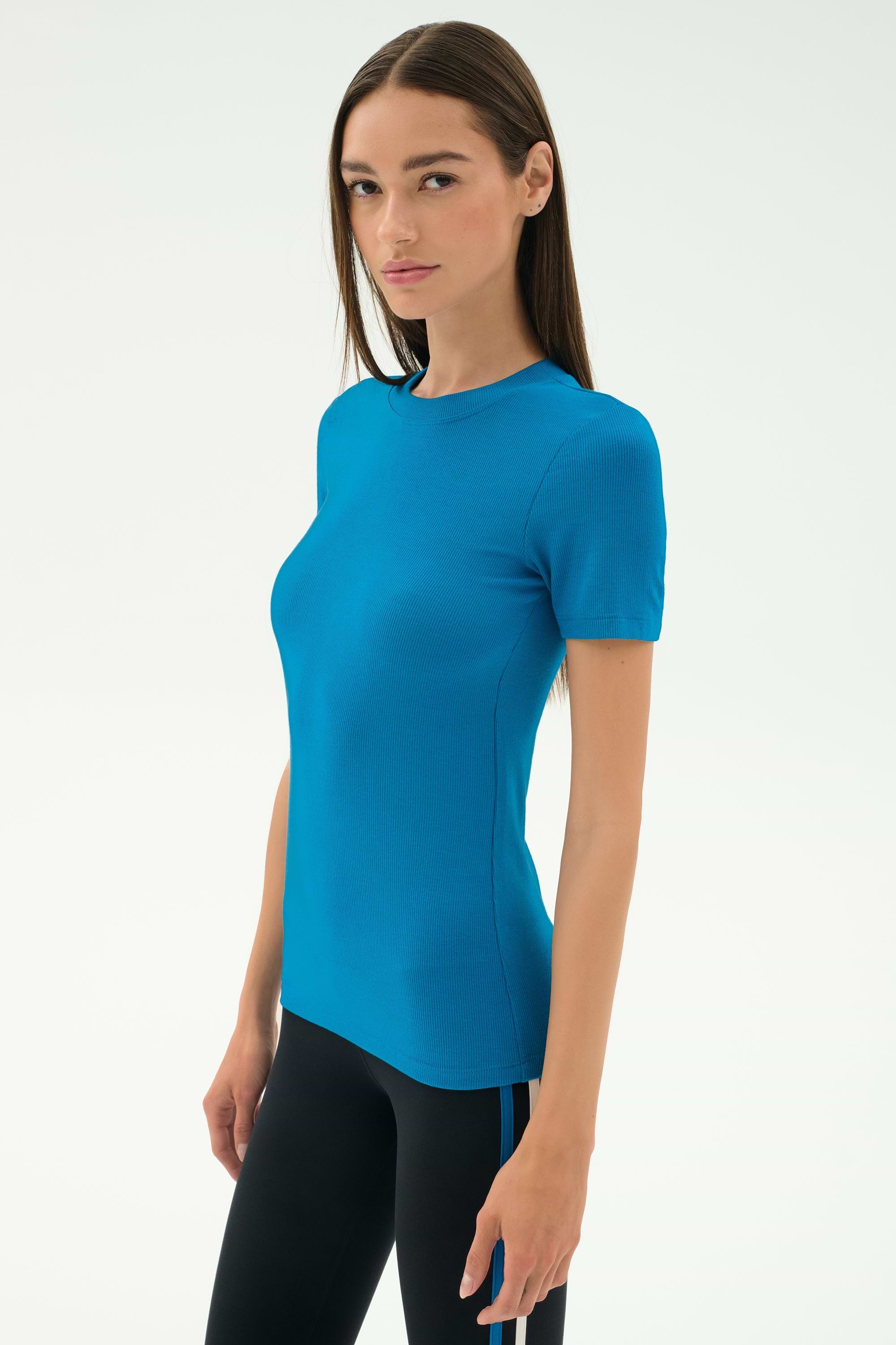 A woman with straight brown hair wears the SPLITS59 Louise Rib Short Sleeve in Stone Blue and black leggings, standing against a plain white background, ready for yoga or gym workouts.