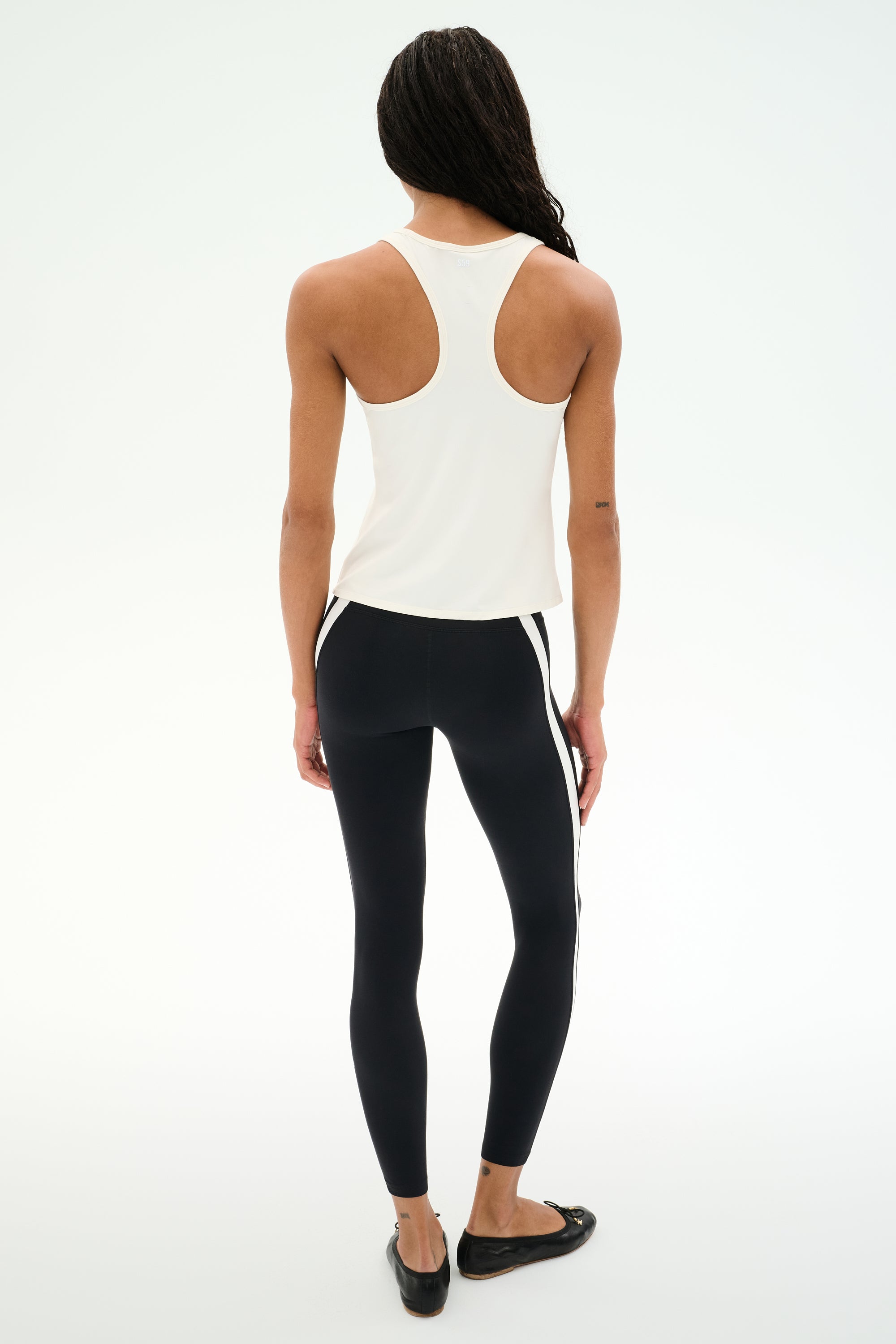A person stands on a white background, facing away, wearing the SPLITS59 Mel Airweight Lite Tank in white—ideal for Pilates—paired with black leggings featuring white side stripes and black flats.