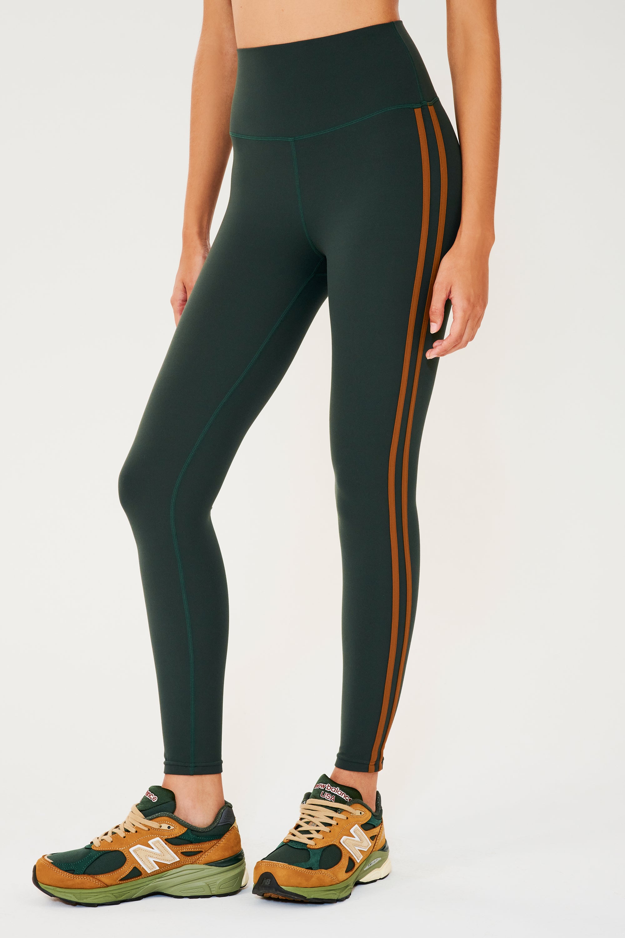 Side view of girl wearing dark green leggings with two thin brown stripes down the side with multi colored shoes