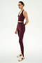 A person stands in profile against a plain white background, wearing the SPLITS59 Rory Airweight Halter Bralette in Dark Cherry/Black, paired with matching stretchy leggings and white Adidas sneakers.