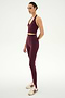 A person stands in a studio setting wearing the SPLITS59 River Dual Waist Airweight Stirrup leggings in Dark Cherry/Black, paired with a matching burgundy sports bra and white sneakers featuring maroon stripes.