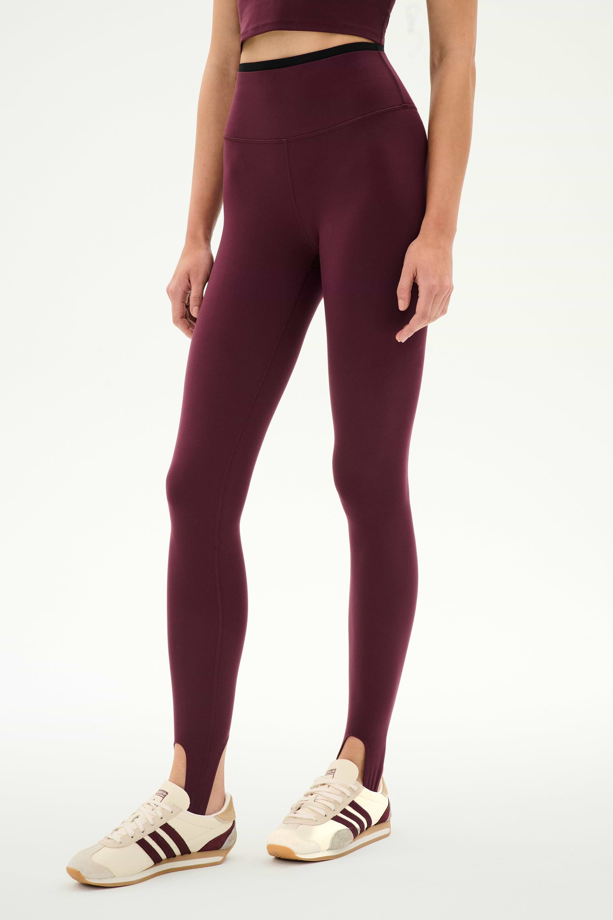 A person wearing the SPLITS59 River Dual Waist Airweight Stirrup leggings in Dark Cherry/Black, featuring a high-waisted fit and stirrup design crafted from smooth nylon spandex fabric, styled with a matching crop top and cream sneakers with maroon stripes, stands against a plain white background.