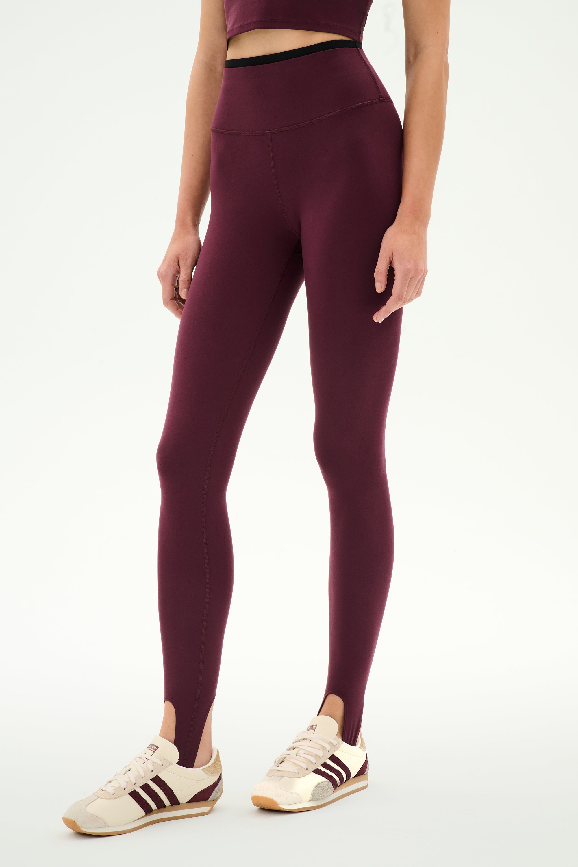 A person wearing the SPLITS59 River Dual Waist Airweight Stirrup leggings in Dark Cherry/Black, featuring a high-waisted fit and stirrup design crafted from smooth nylon spandex fabric, styled with a matching crop top and cream sneakers with maroon stripes, stands against a plain white background.
