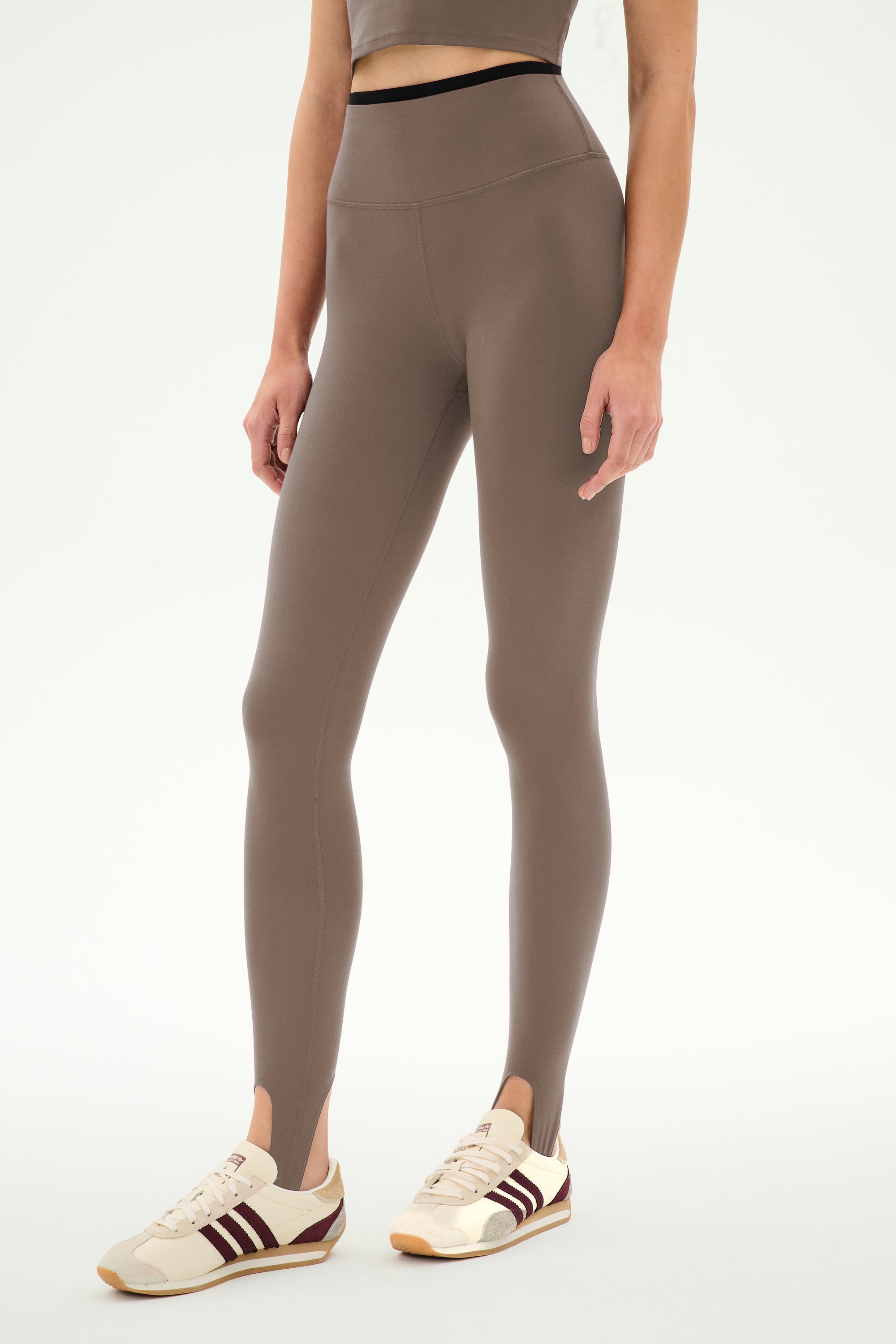 Wearing SPLITS59′s River Dual Waist Airweight Stirrup leggings in Lentil/Black, this individual stands against a plain white background.