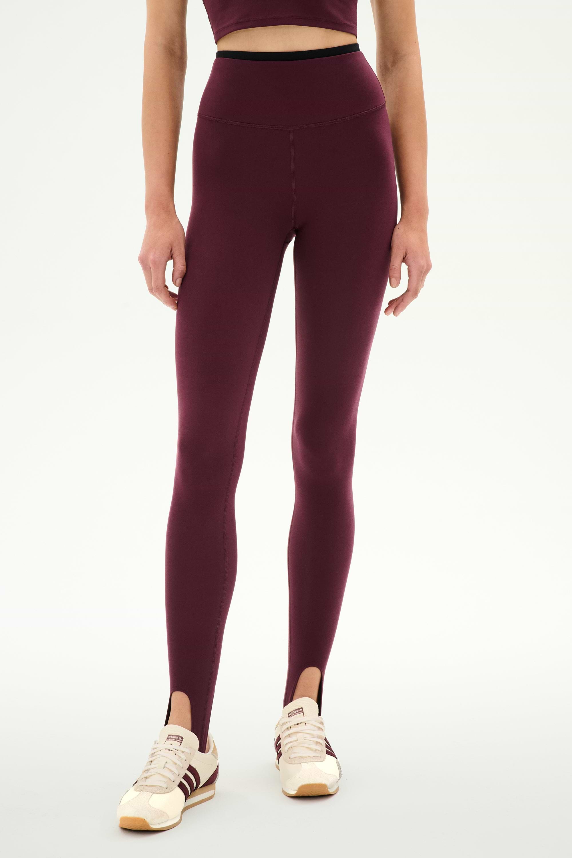 A person wears the SPLITS59 River Dual Waist Airweight Stirrup leggings in Dark Cherry/Black, paired with a matching crop top and white sneakers featuring burgundy stripes, standing against a plain background.