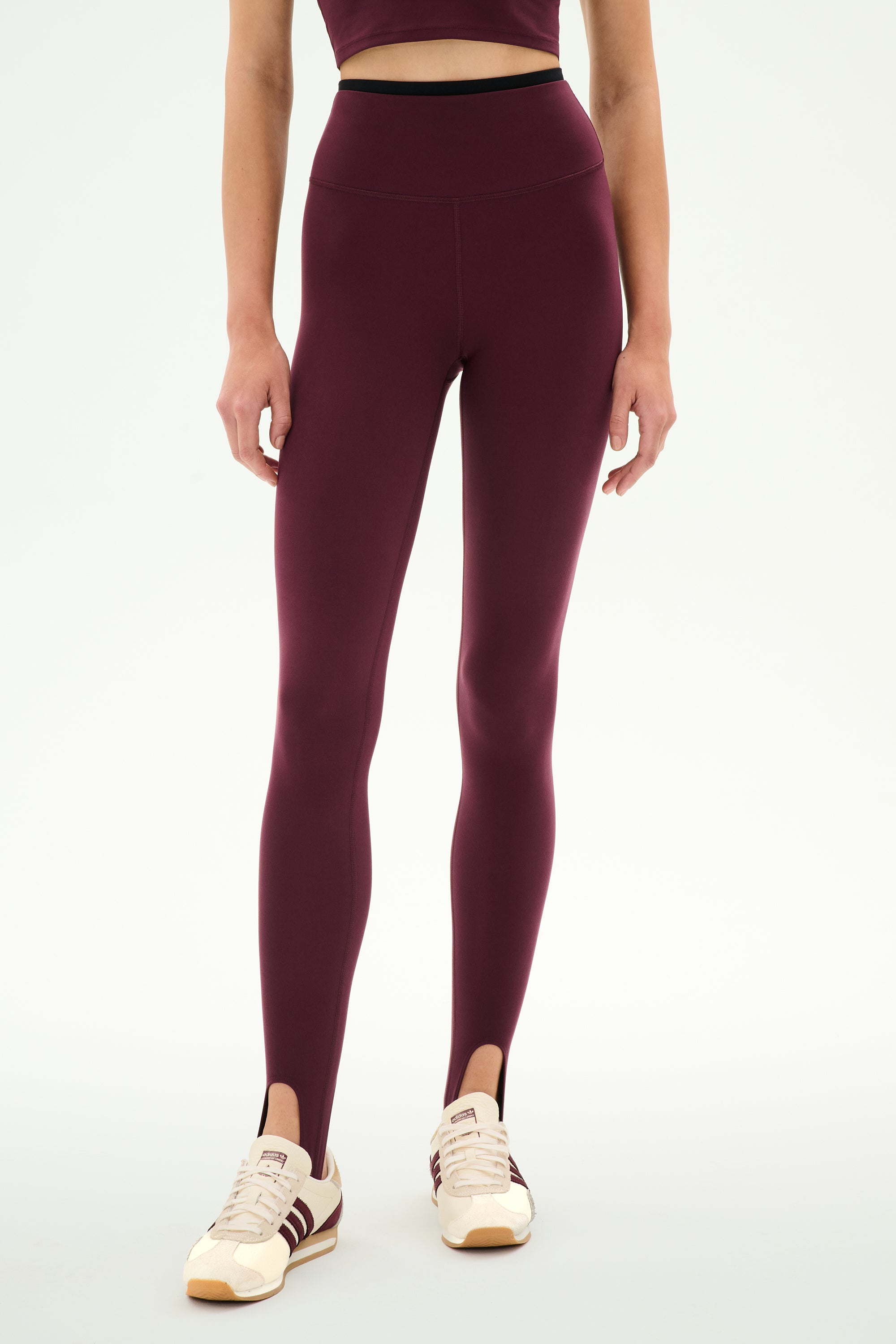 A person wears the SPLITS59 River Dual Waist Airweight Stirrup leggings in Dark Cherry/Black, paired with a matching crop top and white sneakers featuring burgundy stripes, standing against a plain background.