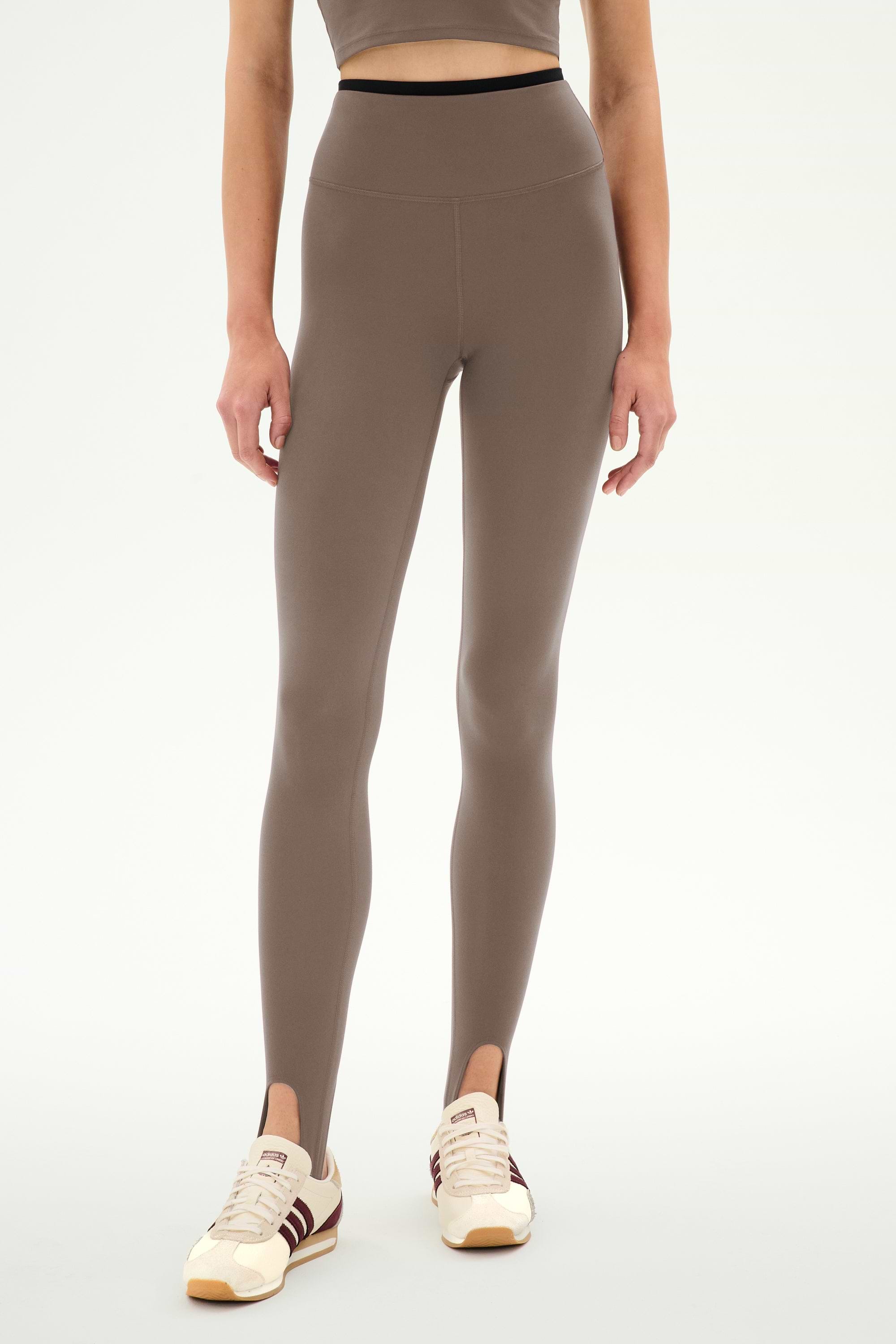 A person is wearing the SPLITS59 River Dual Waist Airweight Stirrup leggings in Lentil/Black, featuring a high-waisted design with stirrups and crafted from a stretchy nylon-spandex blend ideal for athletic wear. The look is paired with a matching crop top and cream sneakers with brown stripes, set against a plain white background.