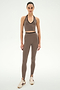 A woman stands facing forward against a plain white background, wearing the SPLITS59 River Dual Waist Airweight Stirrup leggings in Lentil/Black—a high-waisted, brown stretch style with stirrups—paired with a matching crop top and beige sneakers for a comfortable, coordinated look.