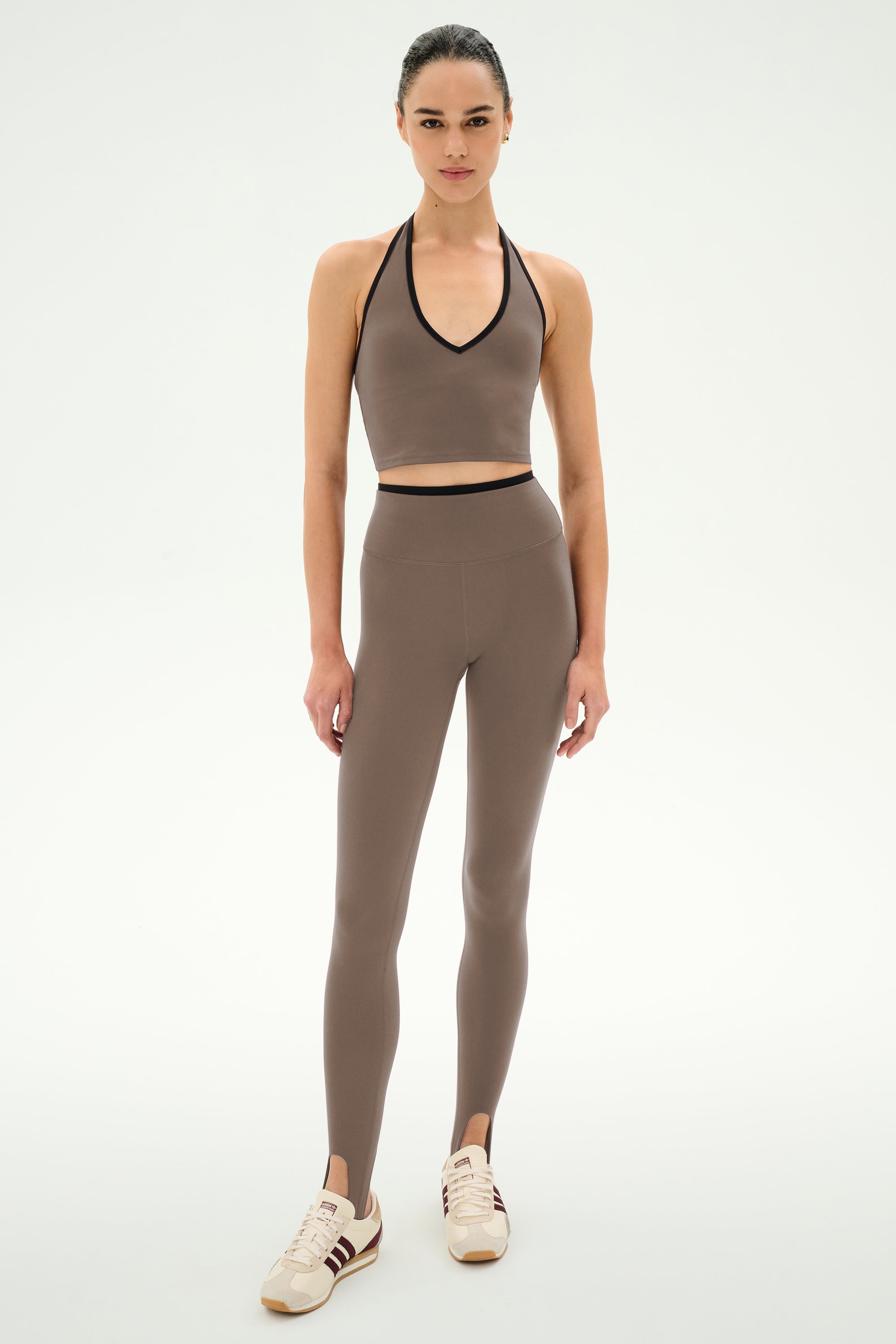 A woman stands facing forward against a plain white background, wearing the SPLITS59 River Dual Waist Airweight Stirrup leggings in Lentil/Black—a high-waisted, brown stretch style with stirrups—paired with a matching crop top and beige sneakers for a comfortable, coordinated look.