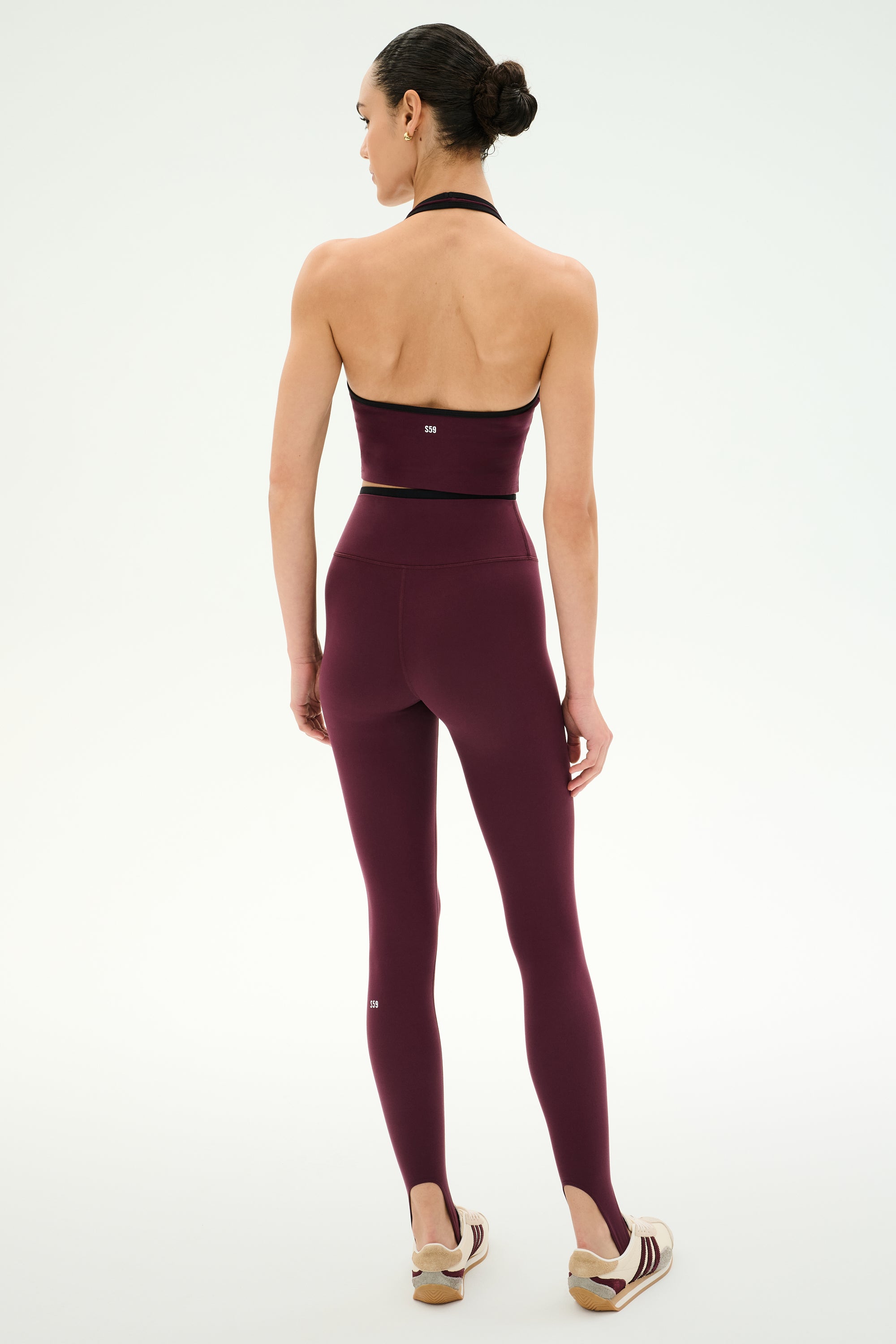 A person stands with their back to the camera, wearing the SPLITS59 Rory Airweight Halter Bralette in Dark Cherry/Black, crafted from stretchy nylon spandex fabric and paired with matching leggings and beige sneakers against a plain white background.
