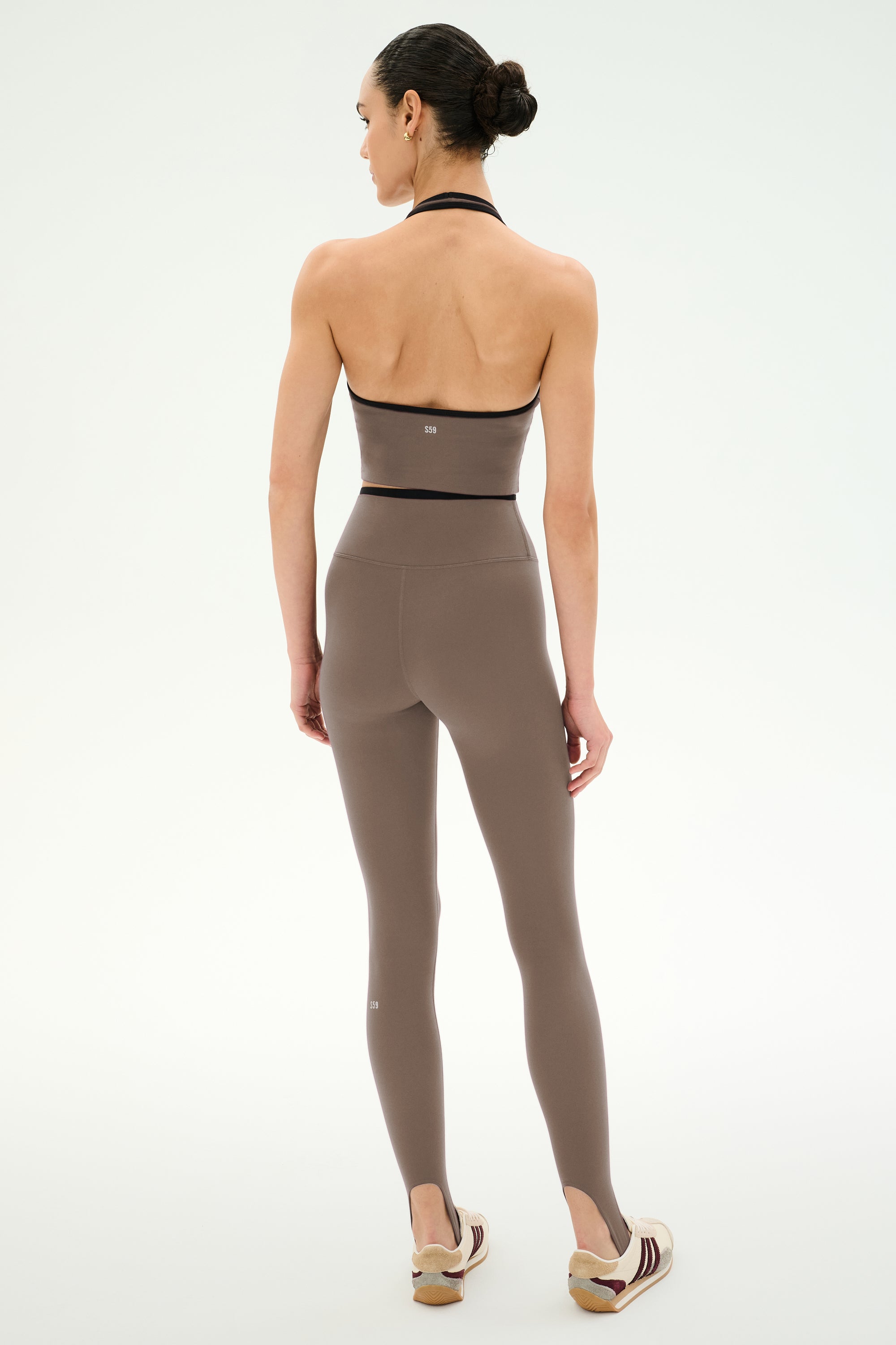 A woman stands facing away, wearing the SPLITS59 River Dual Waist Airweight Stirrup leggings in Lentil/Black, crafted from a sleek nylon-spandex stretch fabric and styled with sneakers, against a plain white background.