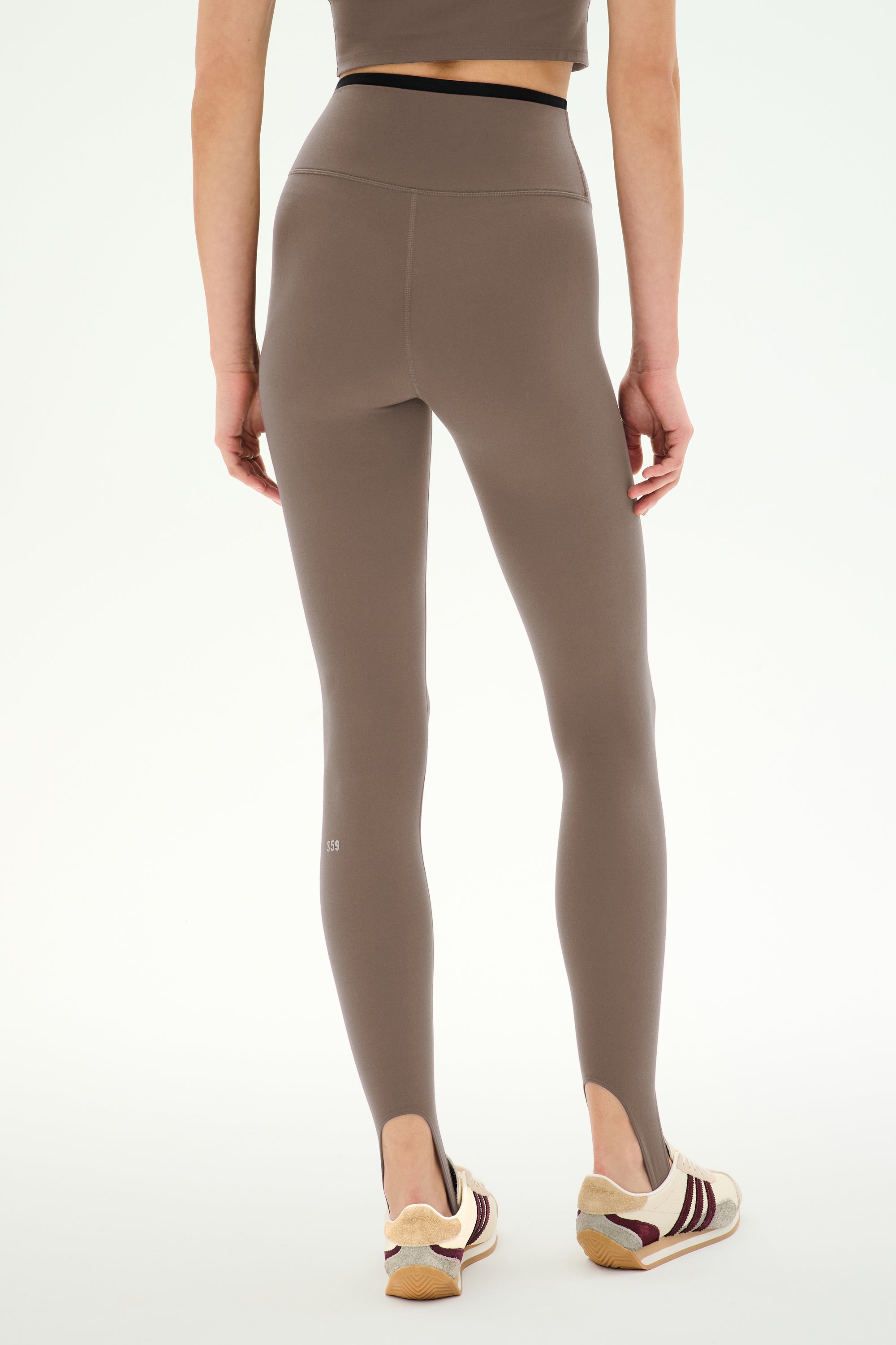 A person stands facing away, styled in SPLITS59’s River Dual Waist Airweight Stirrup leggings in Lentil/Black—a high-waisted design with stirrups crafted from a stretchy nylon-spandex blend—paired with a matching crop top and white sneakers featuring maroon and tan accents, set against a plain background for an effortlessly contemporary athletic look.
