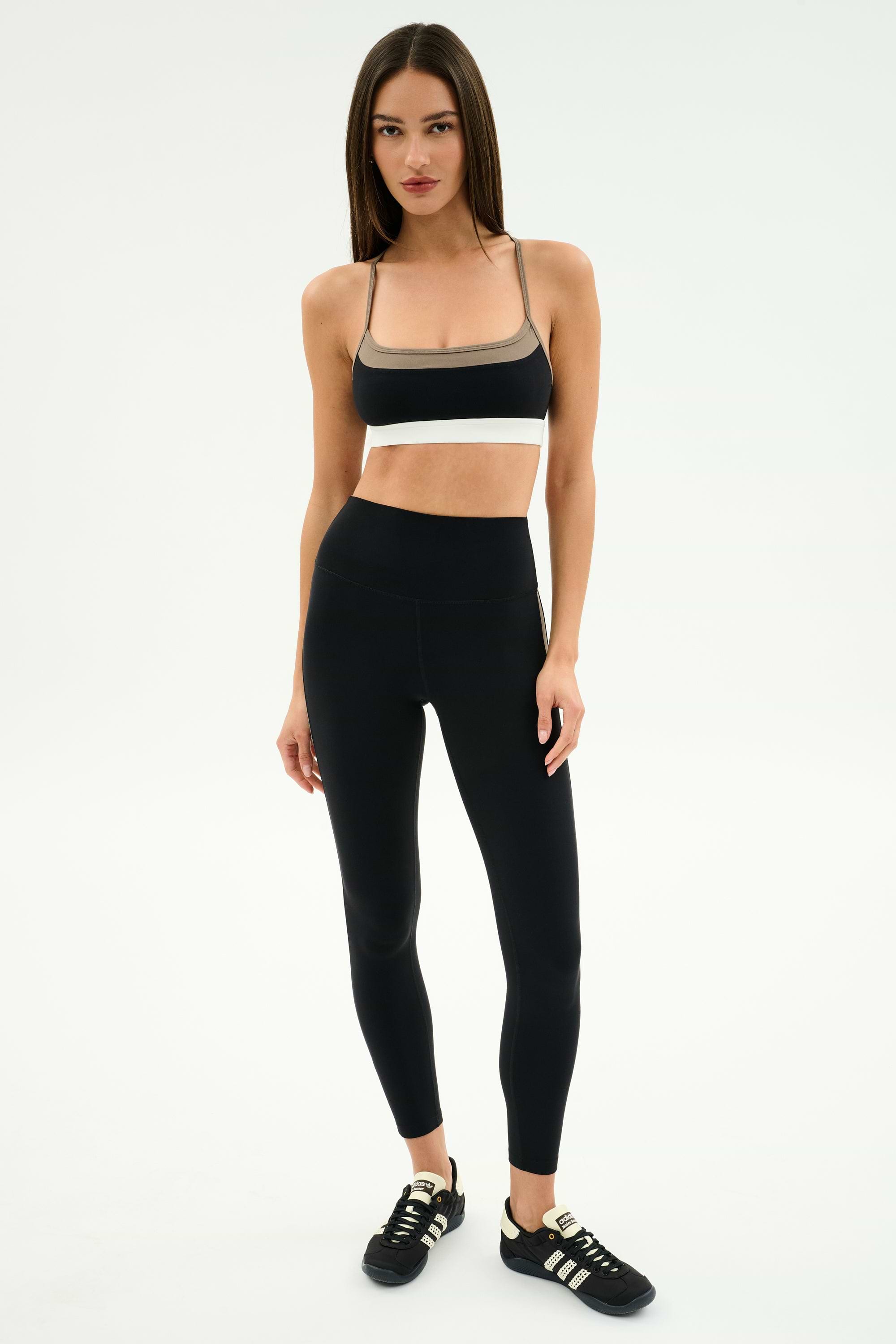 A woman stands against a plain background wearing SPLITS59 Layla Airweight High Waist 7/8 leggings in Black/Lentil with a black sports bra and black Adidas sneakers—ideal attire for hot yoga or gym sessions.