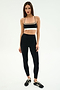 A woman stands against a plain background wearing SPLITS59 Layla Airweight High Waist 7/8 leggings in Black/Lentil with a black sports bra and black Adidas sneakers—ideal attire for hot yoga or gym sessions.