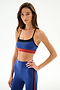 A model wearing a Scottie Airweight Bra in Cobalt/Indigo, paired with matching blue leggings featuring a side stripe and a blue strappy racerback design with black trim and a red hem band.