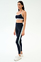 A woman wears the SPLITS59 Emerson Airweight Bra in Indigo/Dove Grey with matching high-waisted leggings, featuring a light blue stripe, styled with white sneakers against a plain light background—ideal for hot yoga.