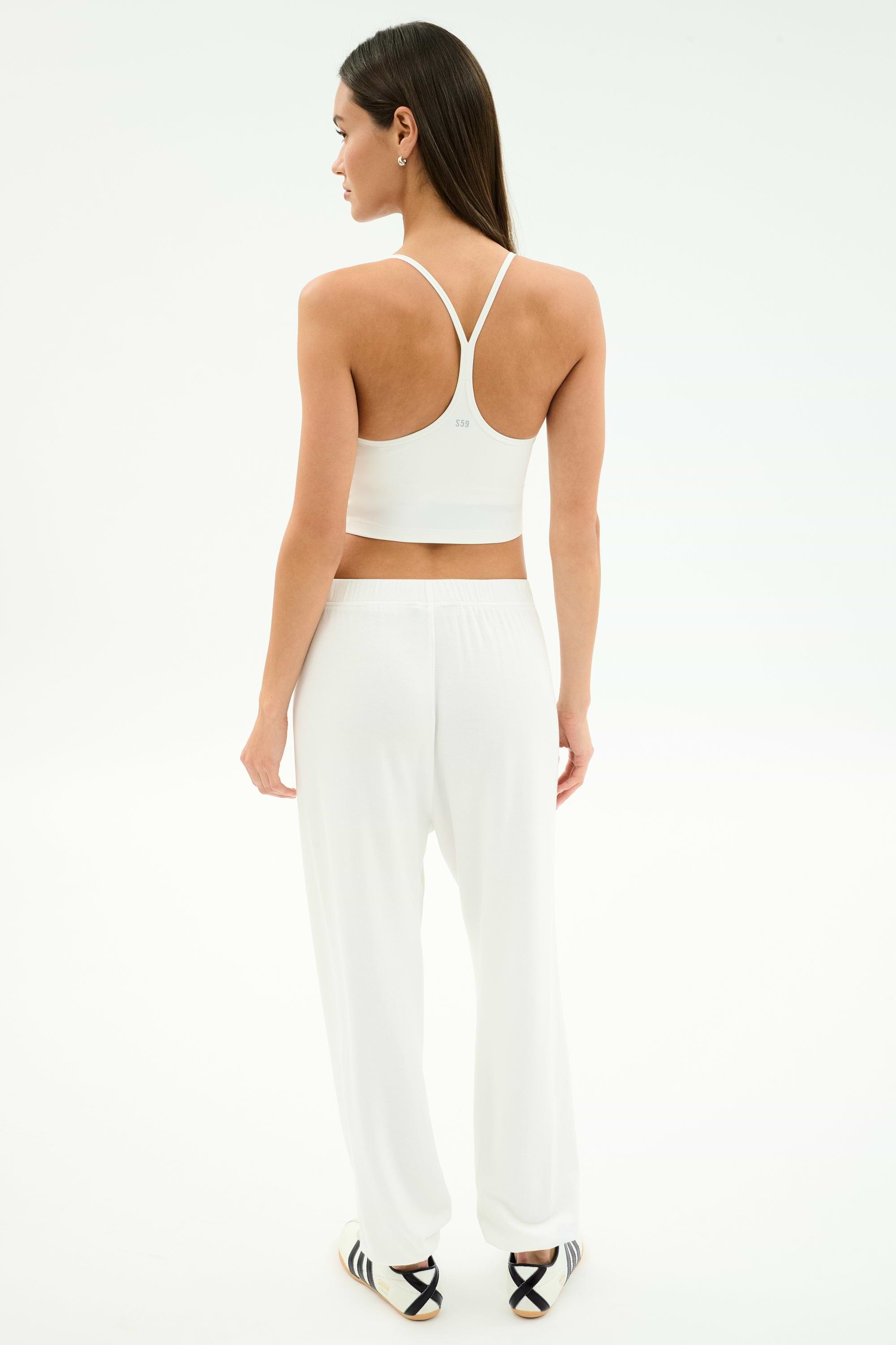 A woman faces away, wearing the SPLITS59 Airweight Crop - White styled as a cropped tank, with loose white pants and striped slide sandals against a plain white background.