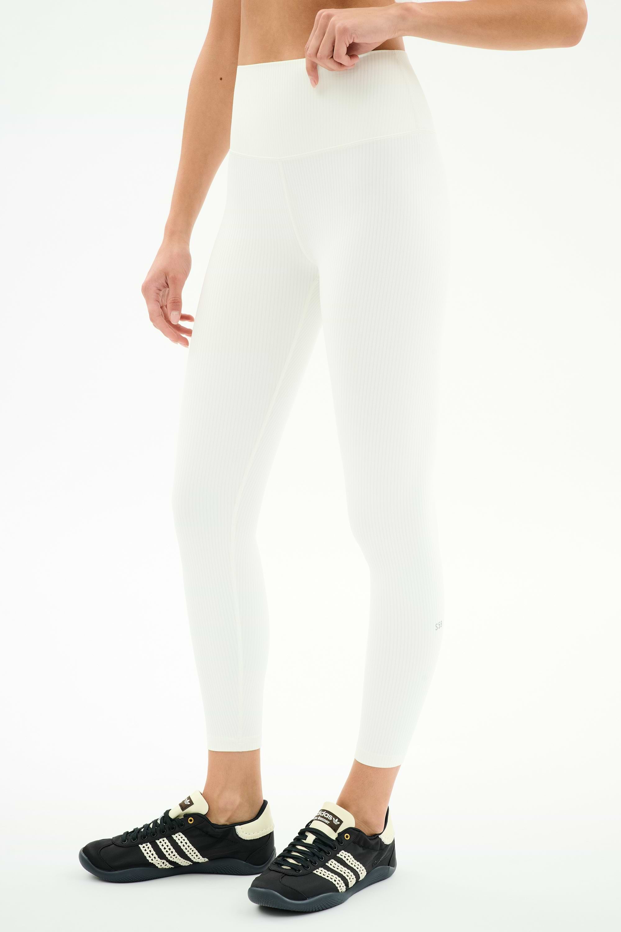 A person wearing SPLITS59 Airweight Rib High Waist 7/8 leggings in white and black Adidas sneakers stands against a plain white background.