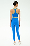 A woman, seen from behind, wears the SPLITS59 Amber Airweight Bralette in Classic Blue/Ray with yellow trim and white sneakers—ideal for hot yoga—set against a plain white background.