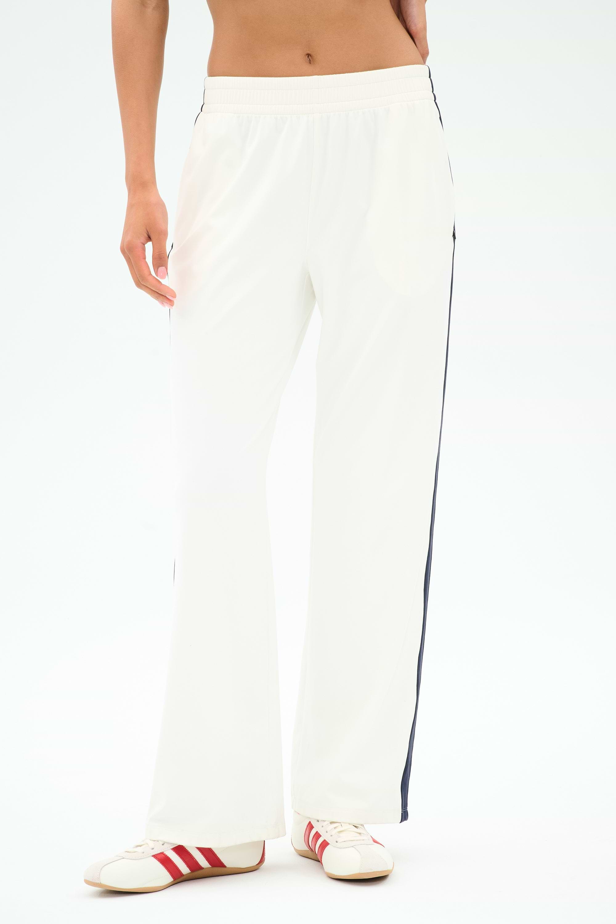 A person wears the SPLITS59 Maxie Rigor Track Pant in White/Indigo with black side stripes and white sneakers with red stripes, standing against a plain light background. Made from soft Rigor fabric for comfort and style.