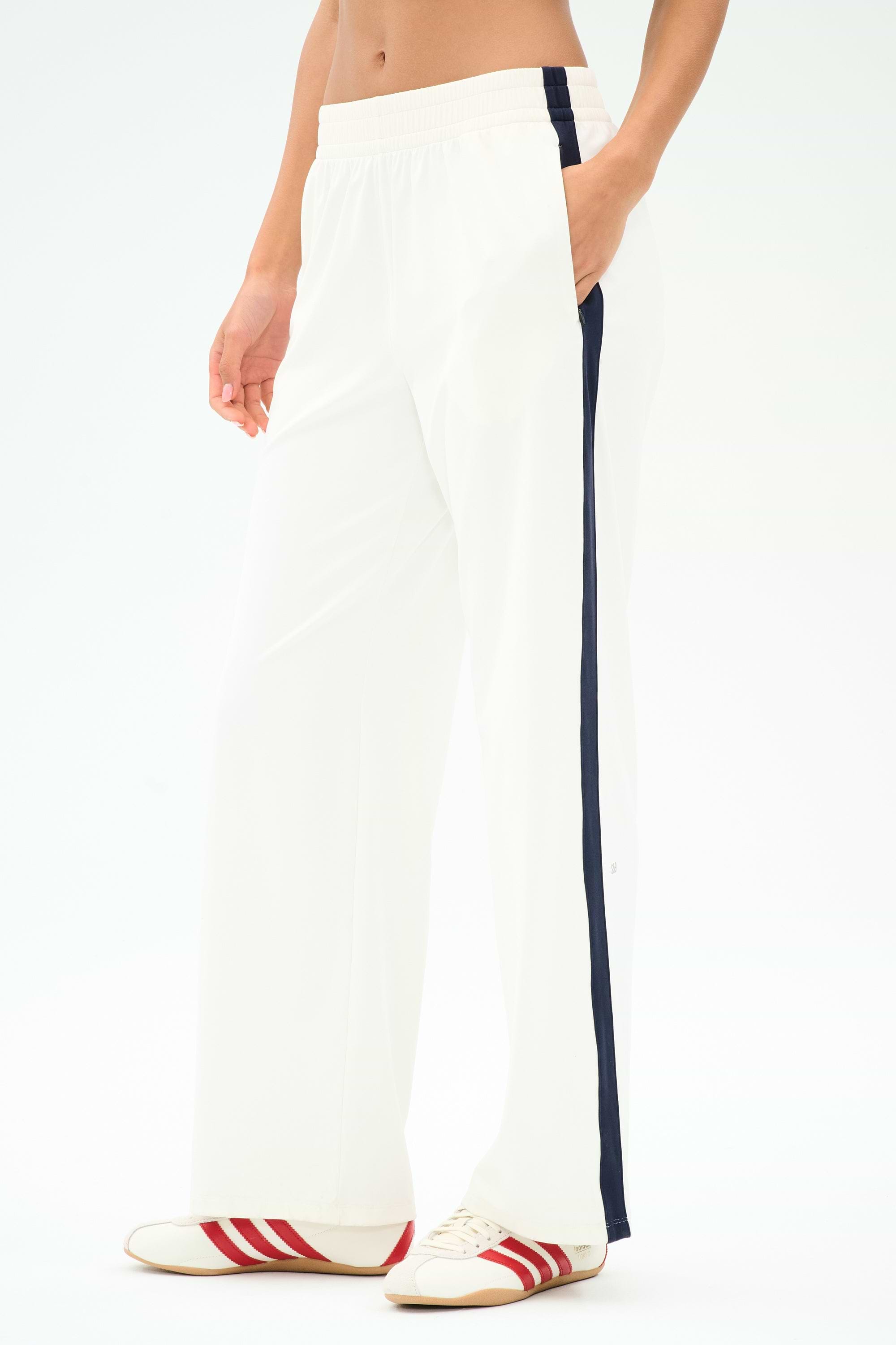 A person wears SPLITS59 Maxie Rigor Track Pant in White/Indigo, featuring a navy side stripe, paired with white sneakers with red stripes, standing against a plain background.