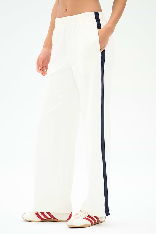 A person wears SPLITS59 Maxie Rigor Track Pant in White/Indigo, featuring a navy side stripe, paired with white sneakers with red stripes, standing against a plain background.